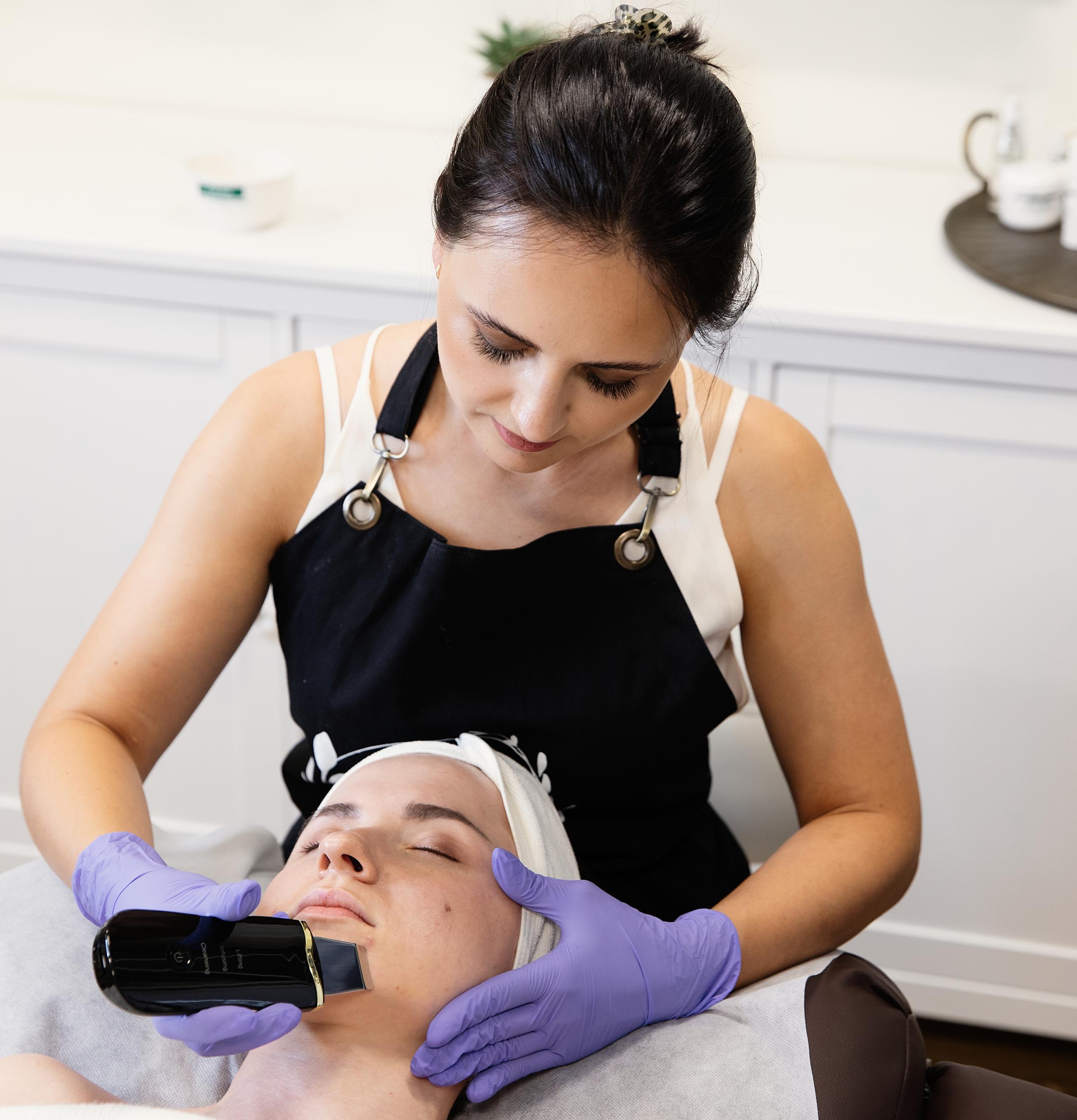 An esthetician in purple gloves uses a black skin scrubber on a client's face.