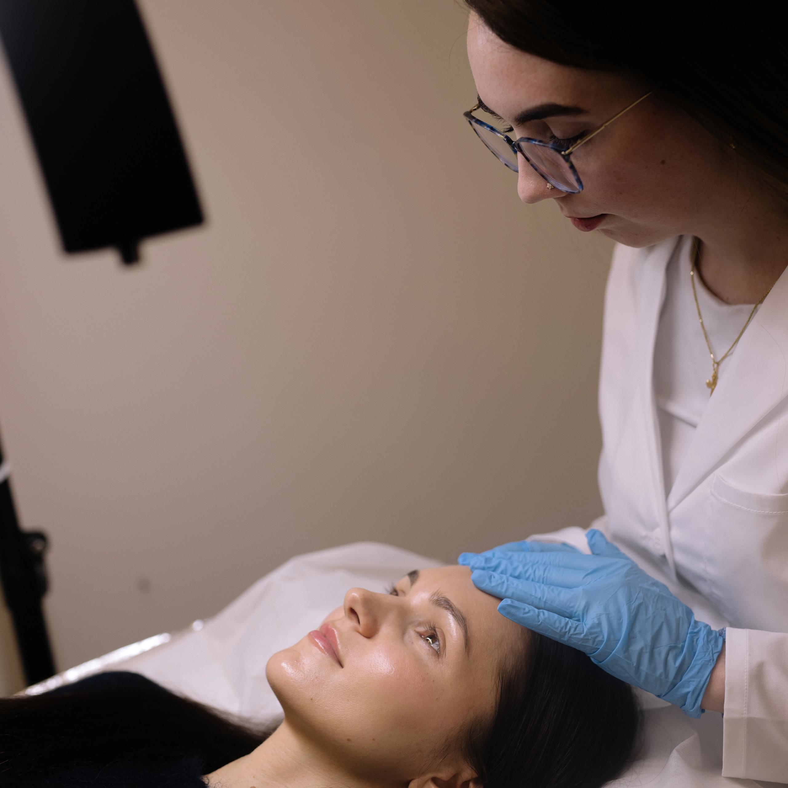 A woman in a lab coat and blue gloves performs a facial treatment on a client.