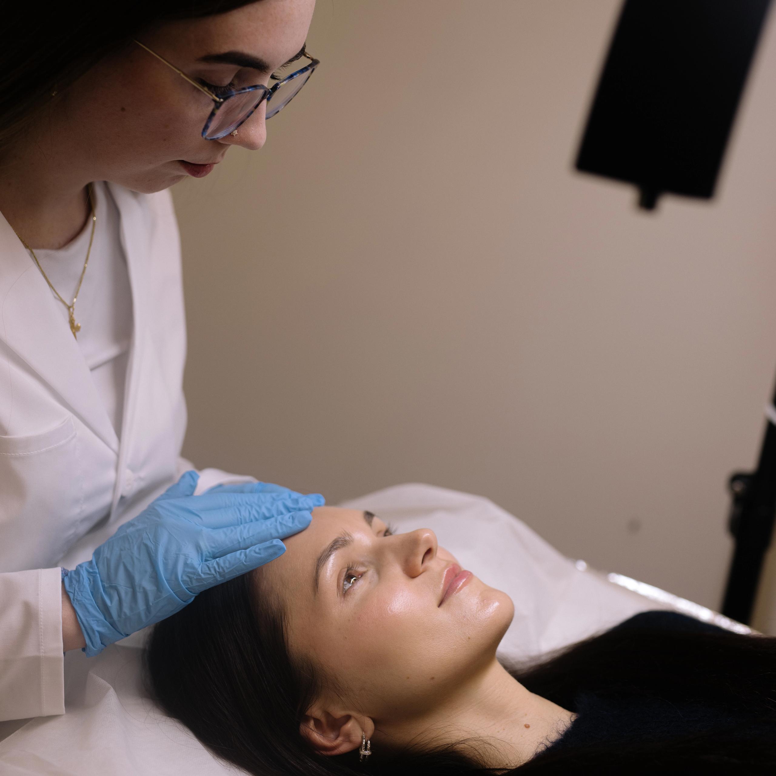 A woman in a white lab coat and blue gloves applies a treatment to a client's forehead.