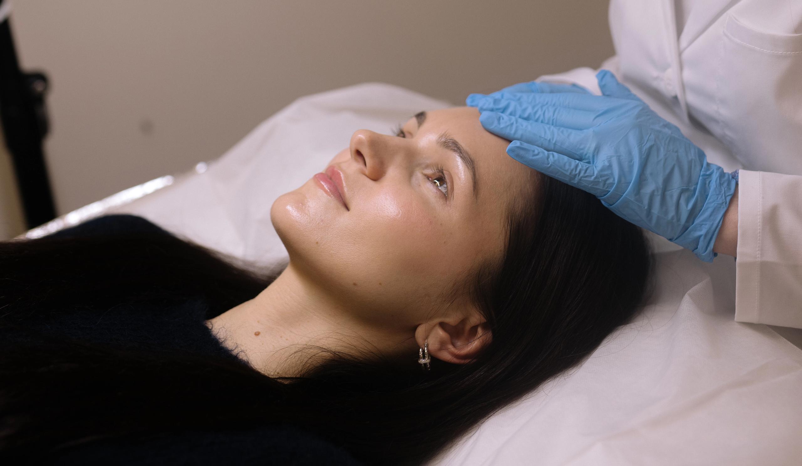 A woman lies down while a gloved hand touches her forehead.
