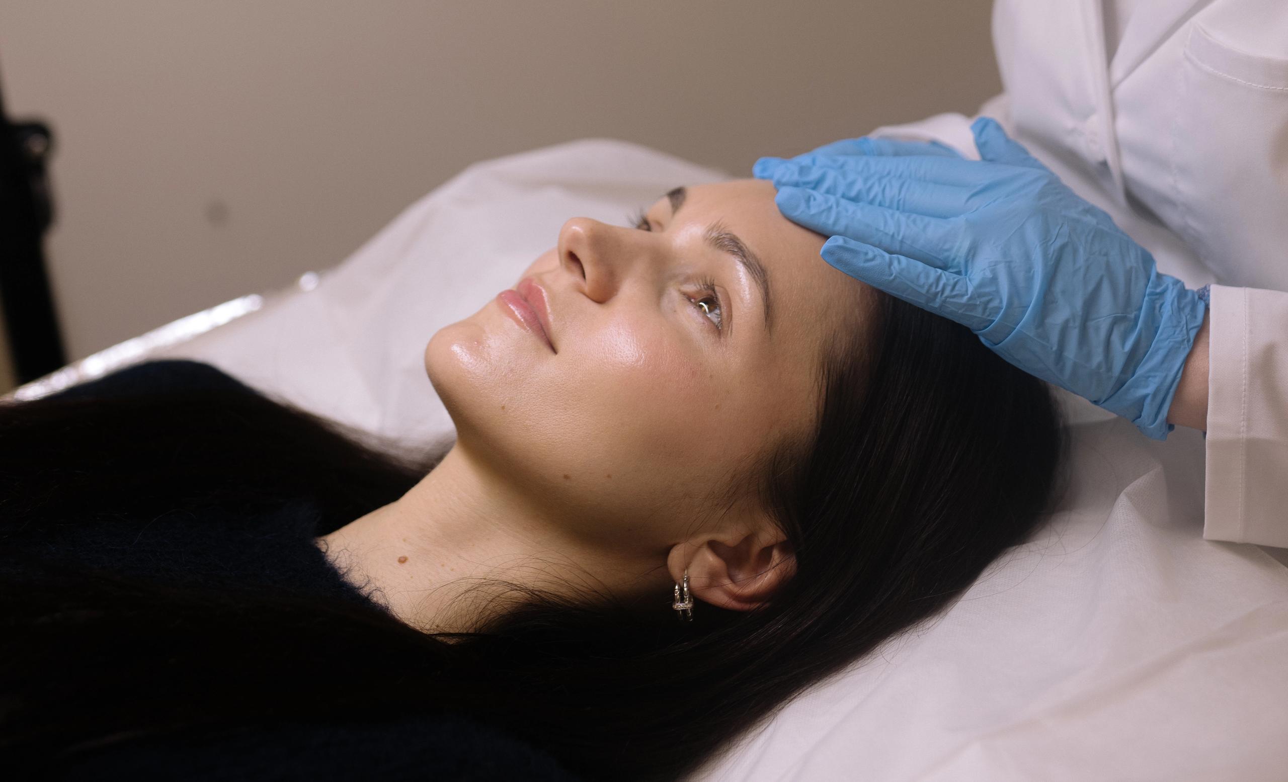 A woman lies down while a gloved hand touches her forehead.