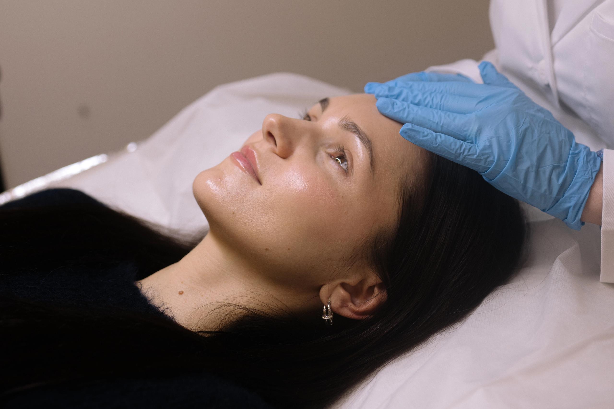 A woman lies down while a gloved hand touches her forehead.