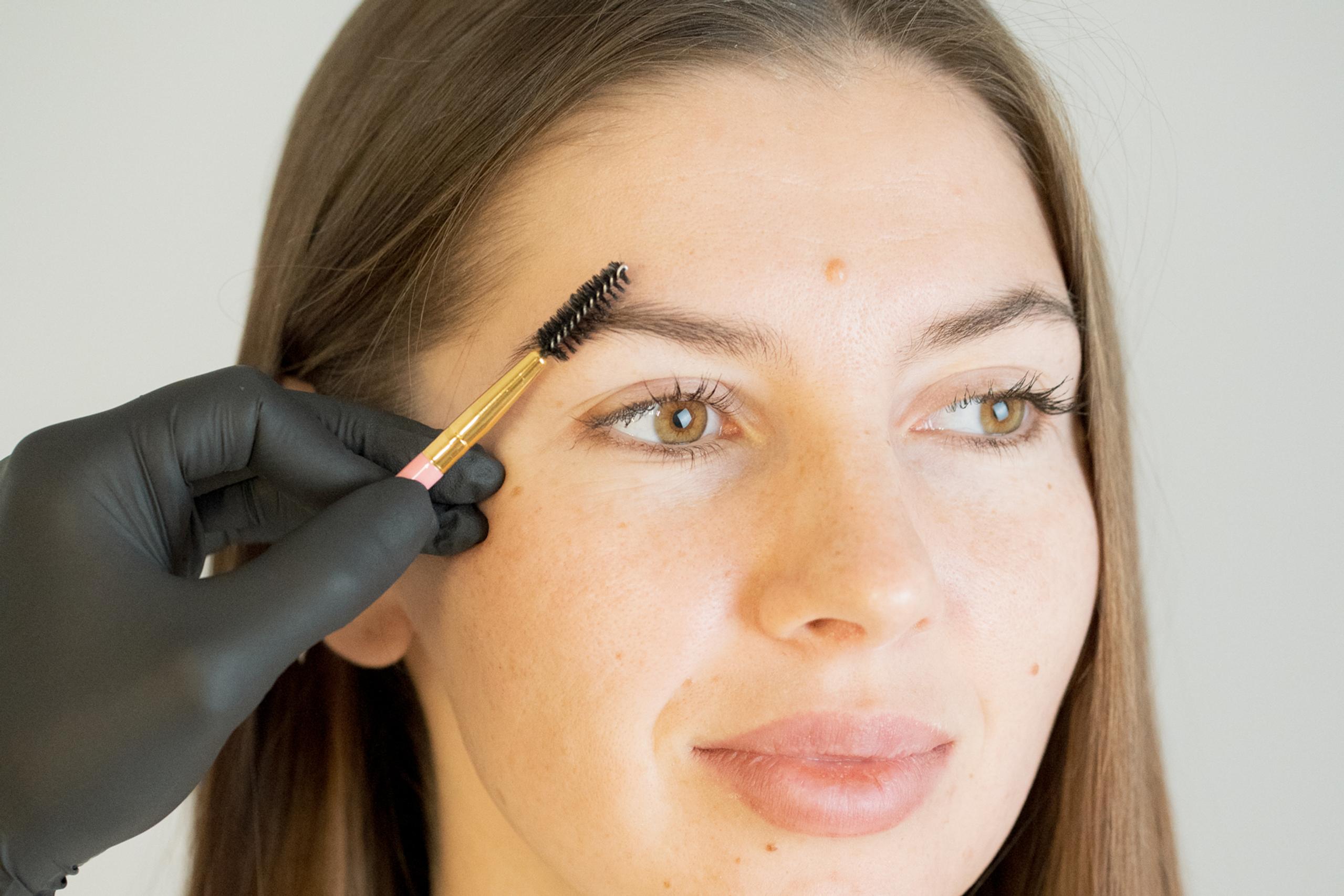 A gloved hand brushes a woman's eyebrow with a spoolie brush.