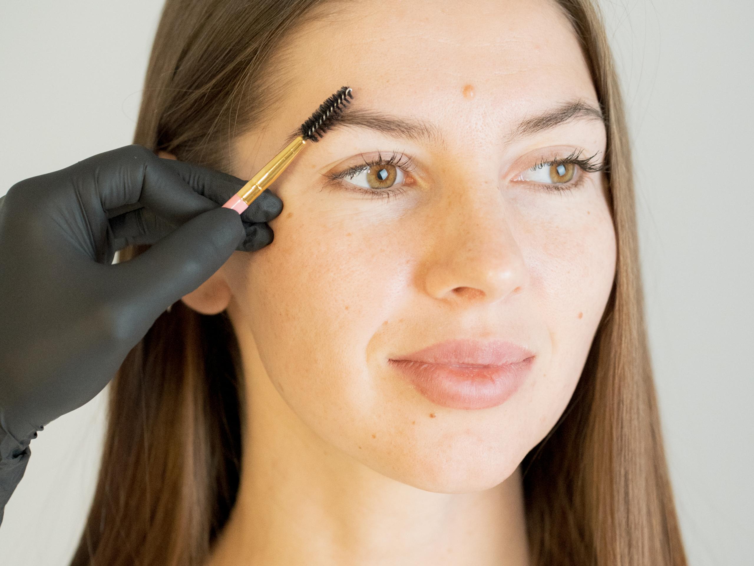 A gloved hand brushes a woman's eyebrow with a spoolie brush.
