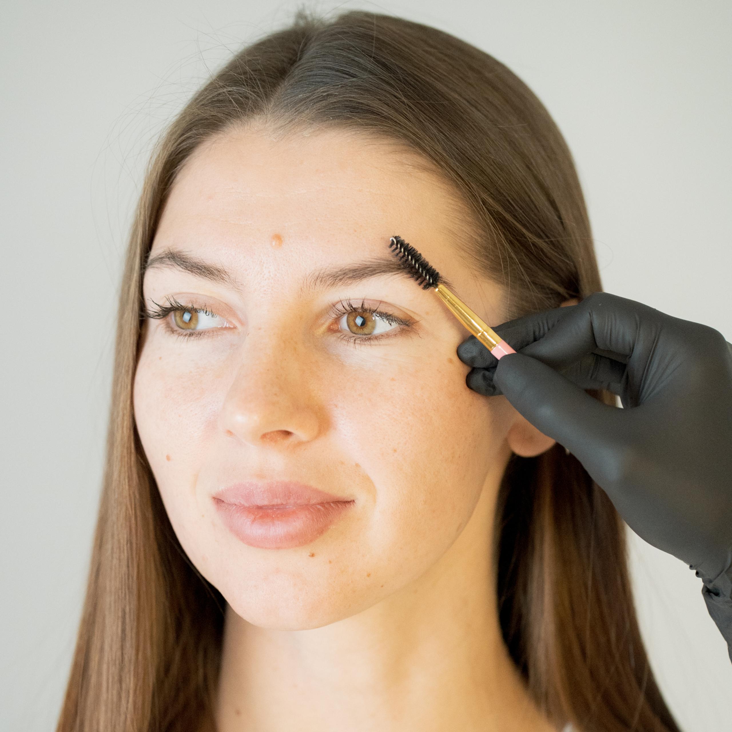 A gloved hand brushes a woman's eyebrow with a spoolie brush.