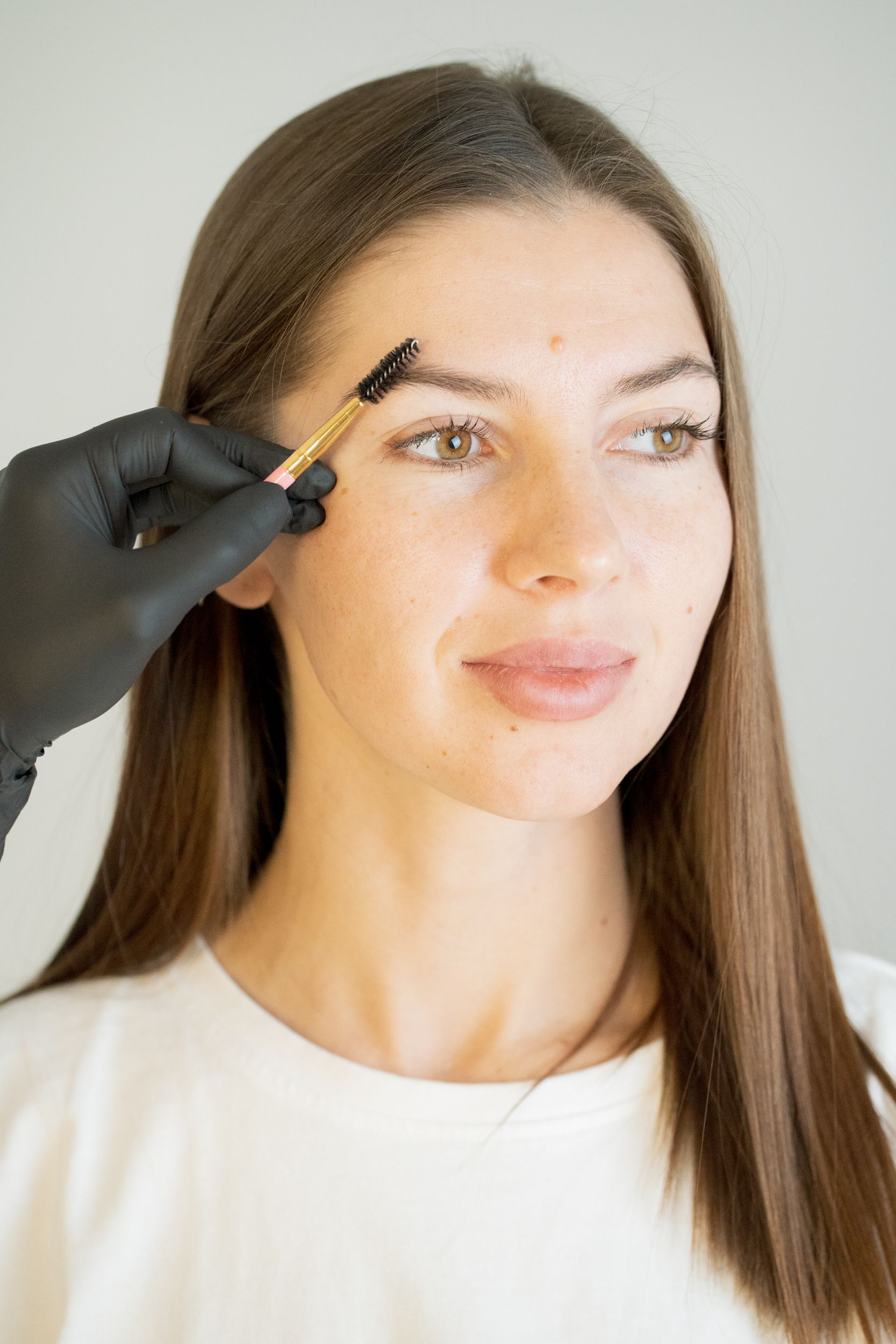 A gloved hand brushes a woman's eyebrow with a spoolie.