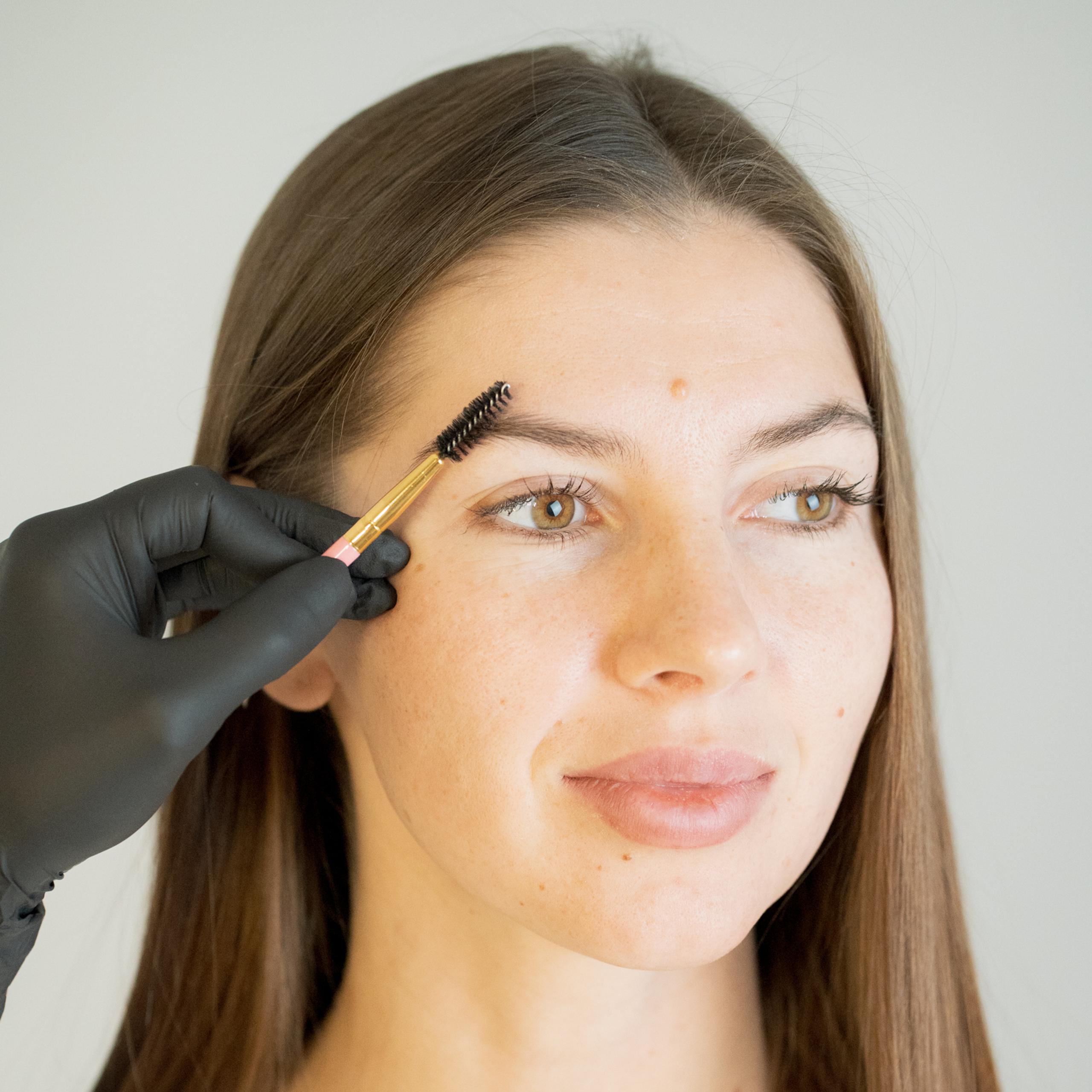 A gloved hand brushes a woman's eyebrow with a spoolie.