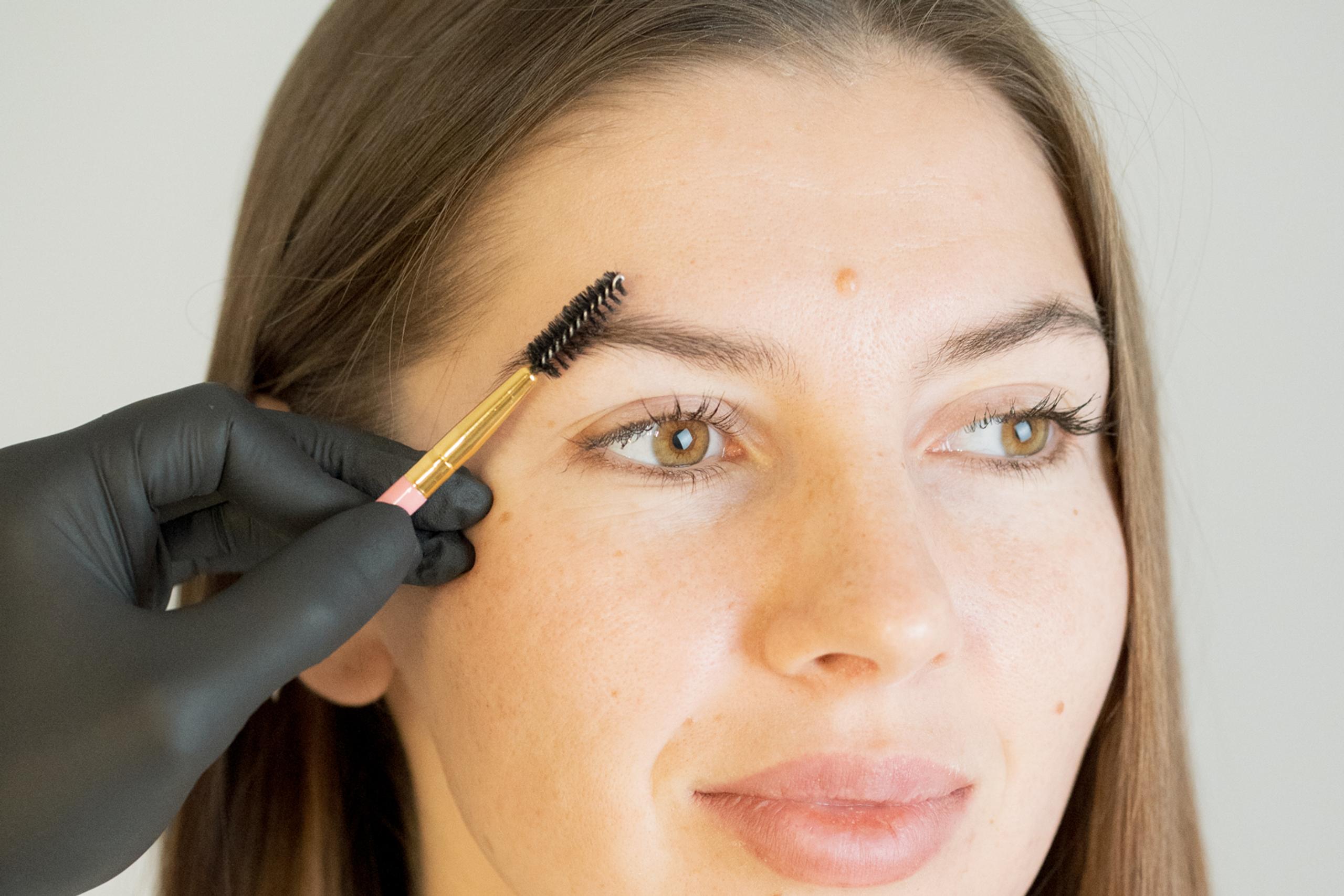A gloved hand brushes a woman's eyebrow with a spoolie.