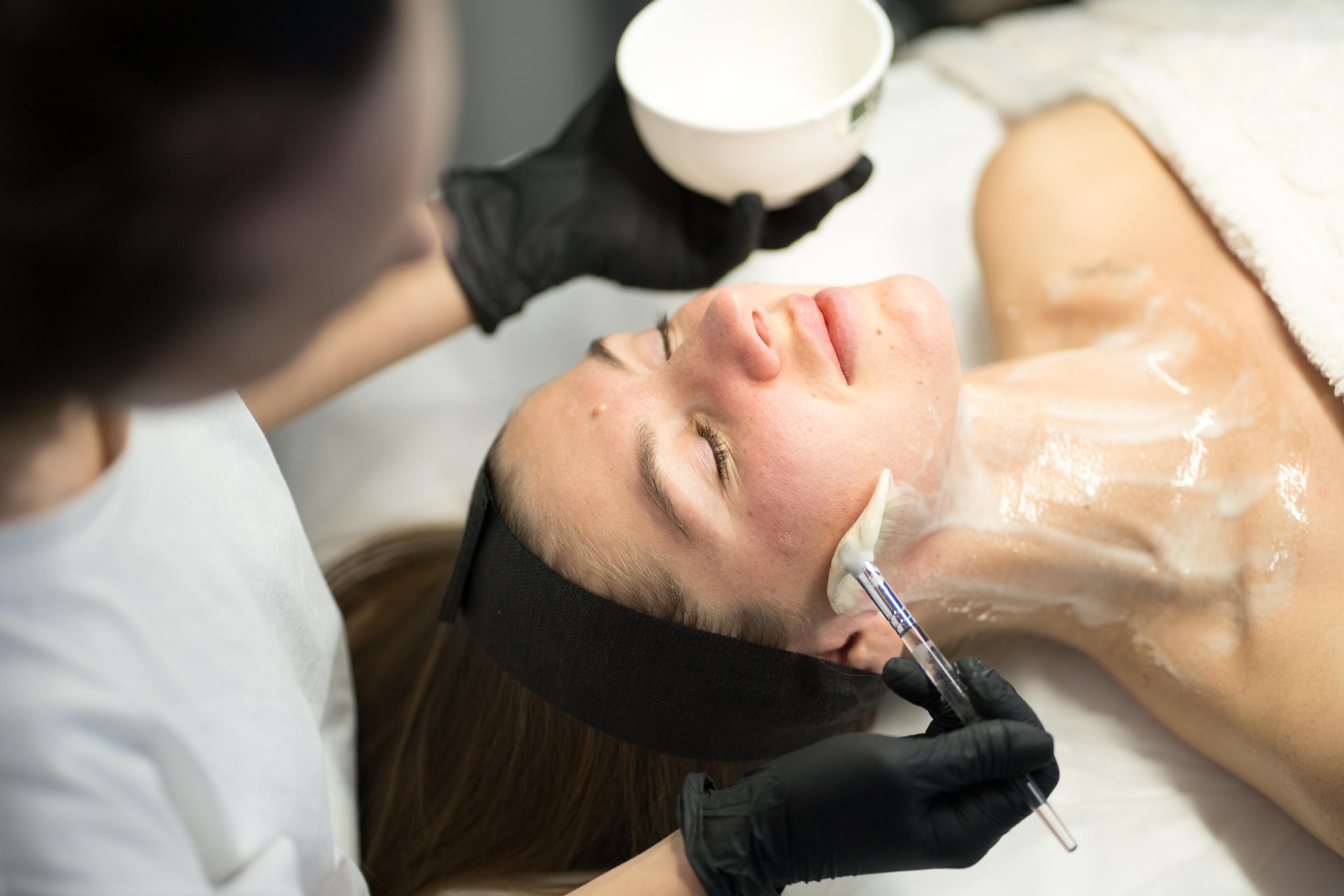 A skincare professional applies a white mask to a client's face and neck with a brush.