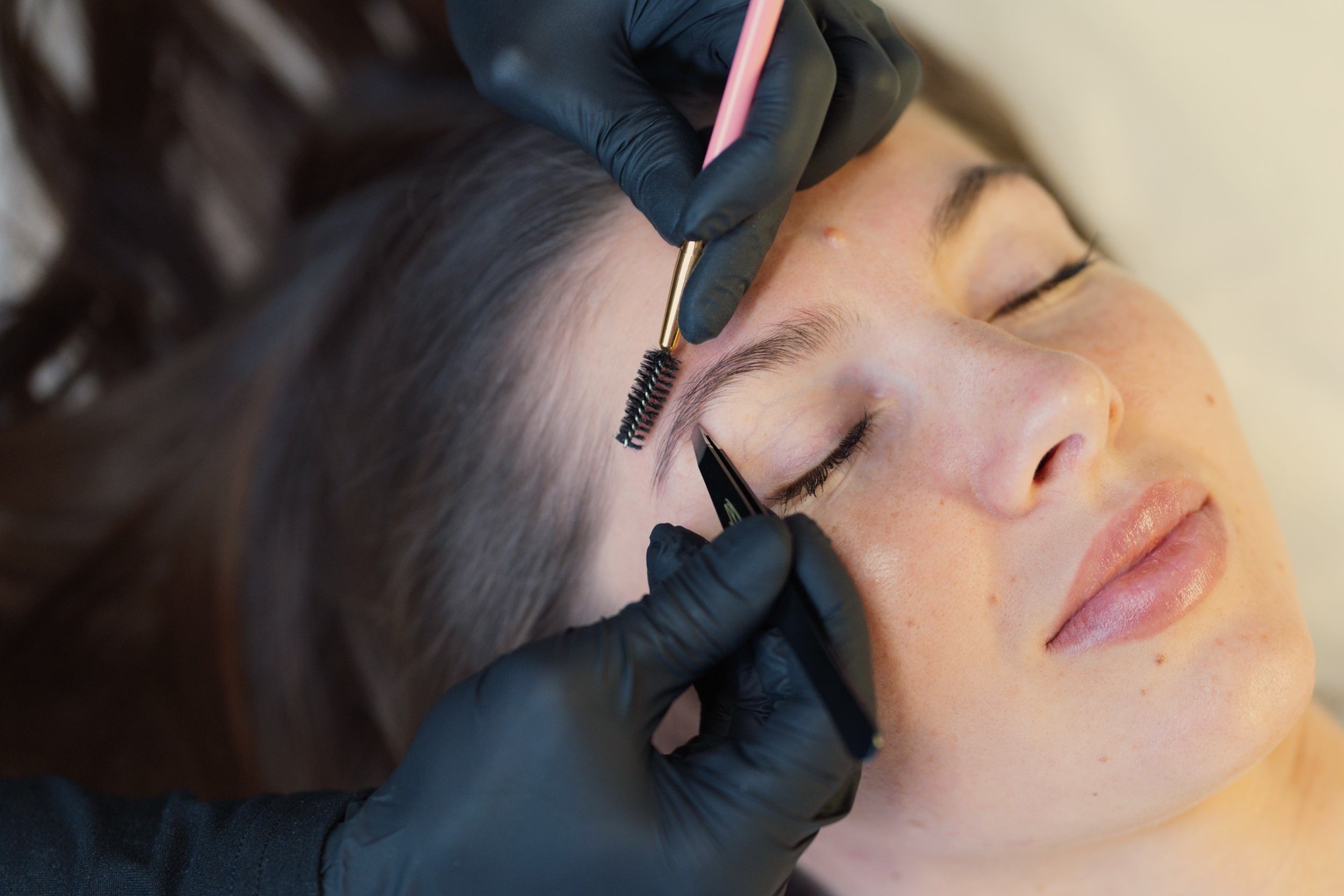 Person in black gloves grooming a woman's eyebrow with a spoolie brush and precision tool.