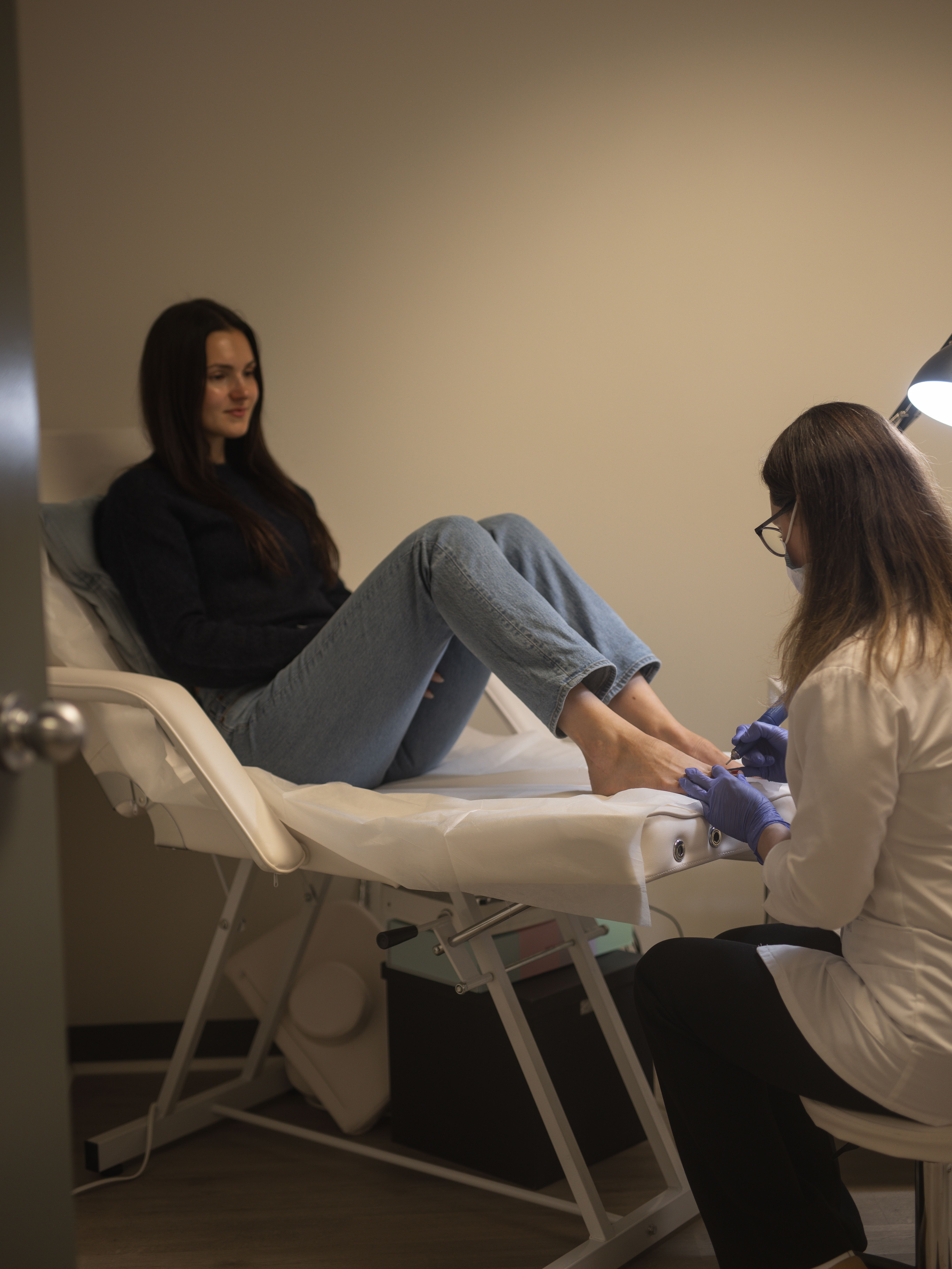 A smiling woman lies on a bed as a technician gives her a pedicure.