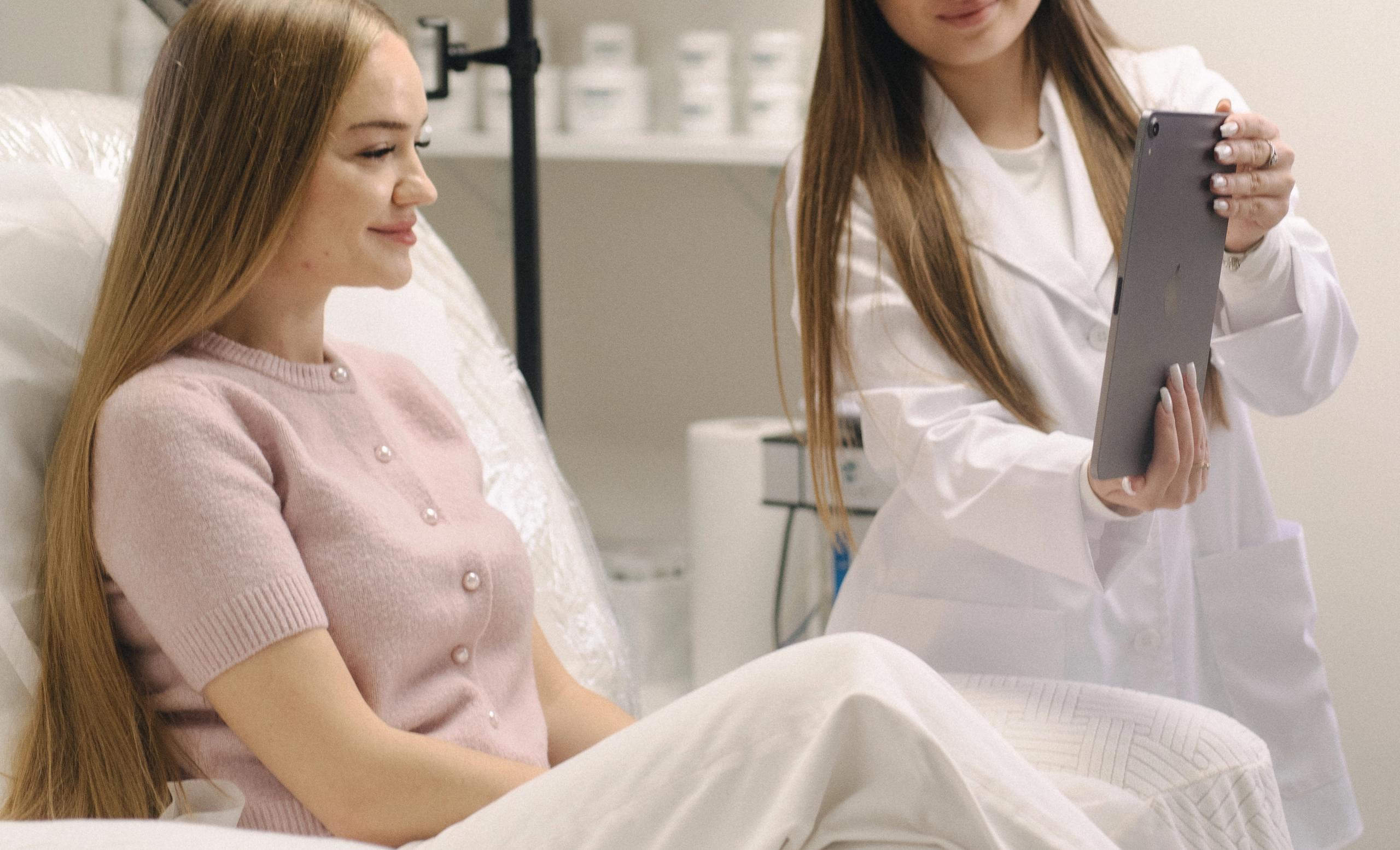 An esthetician shows a tablet to a smiling client seated in a treatment chair.