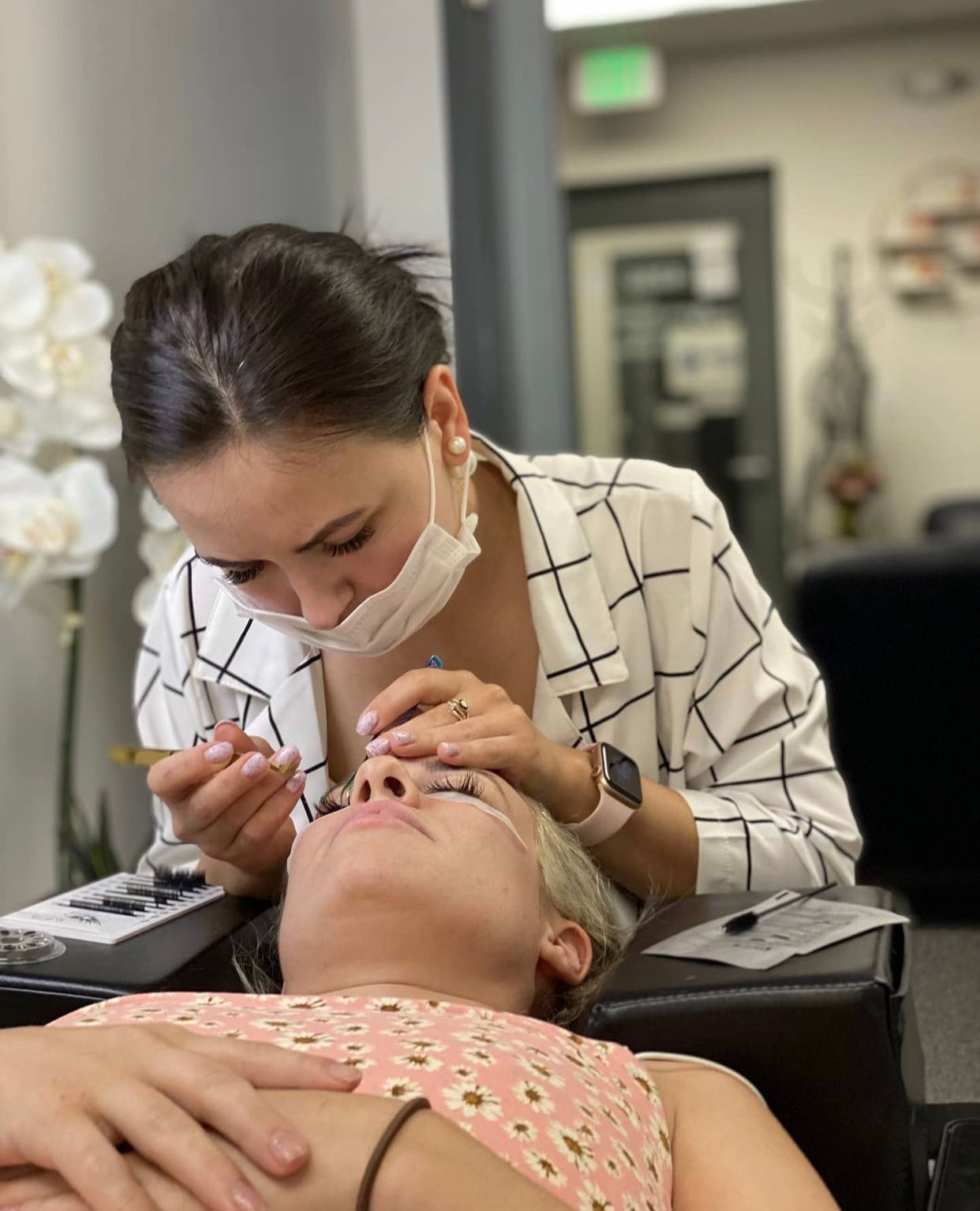 A masked technician applies eyelash extensions to a client.