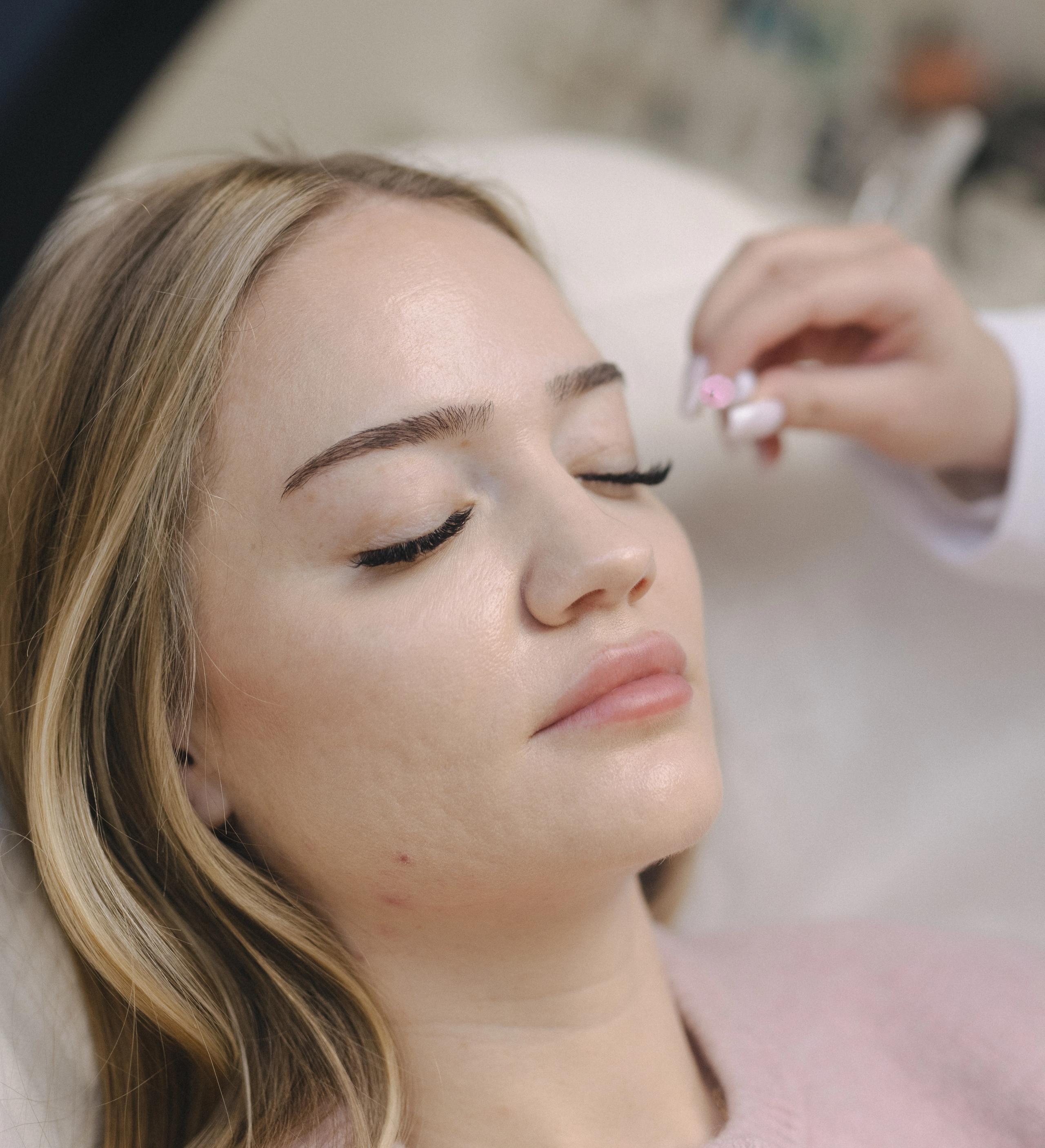 A blonde woman with closed eyes receives a facial treatment.