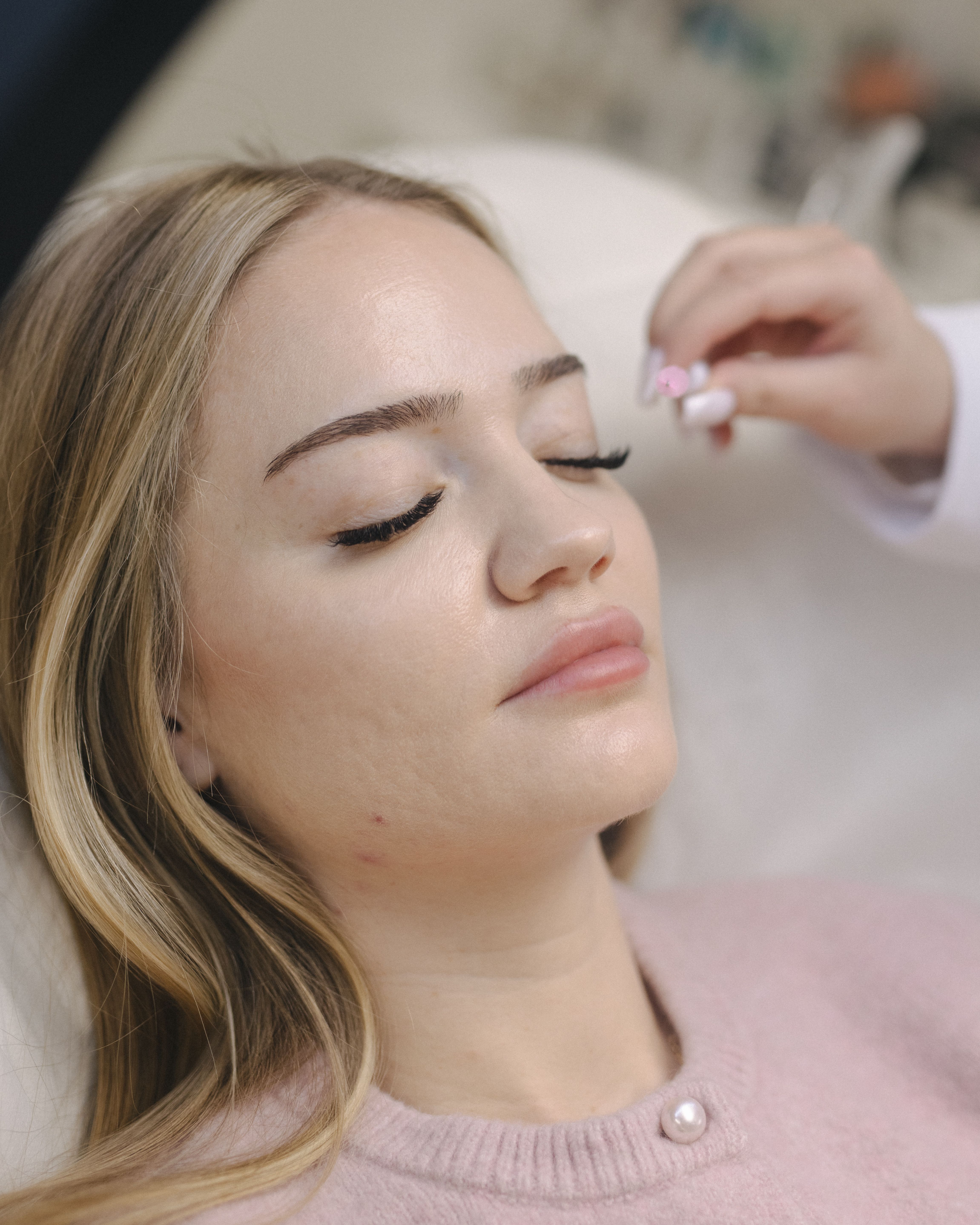 A woman with blonde hair lies with eyes closed as a hand is about to brush her eyelashes