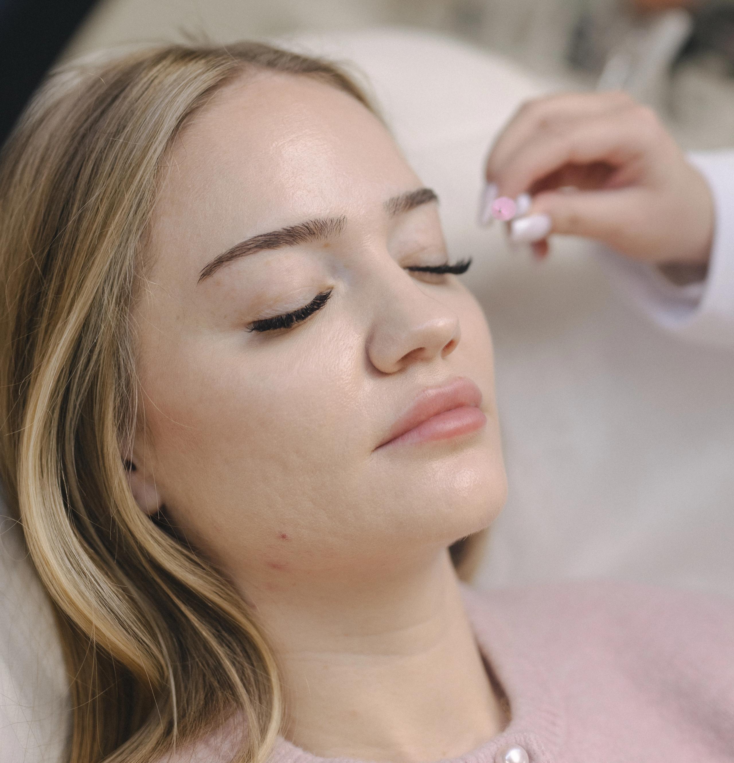A woman with blonde hair lies with eyes closed as a hand is about to brush her eyelashes