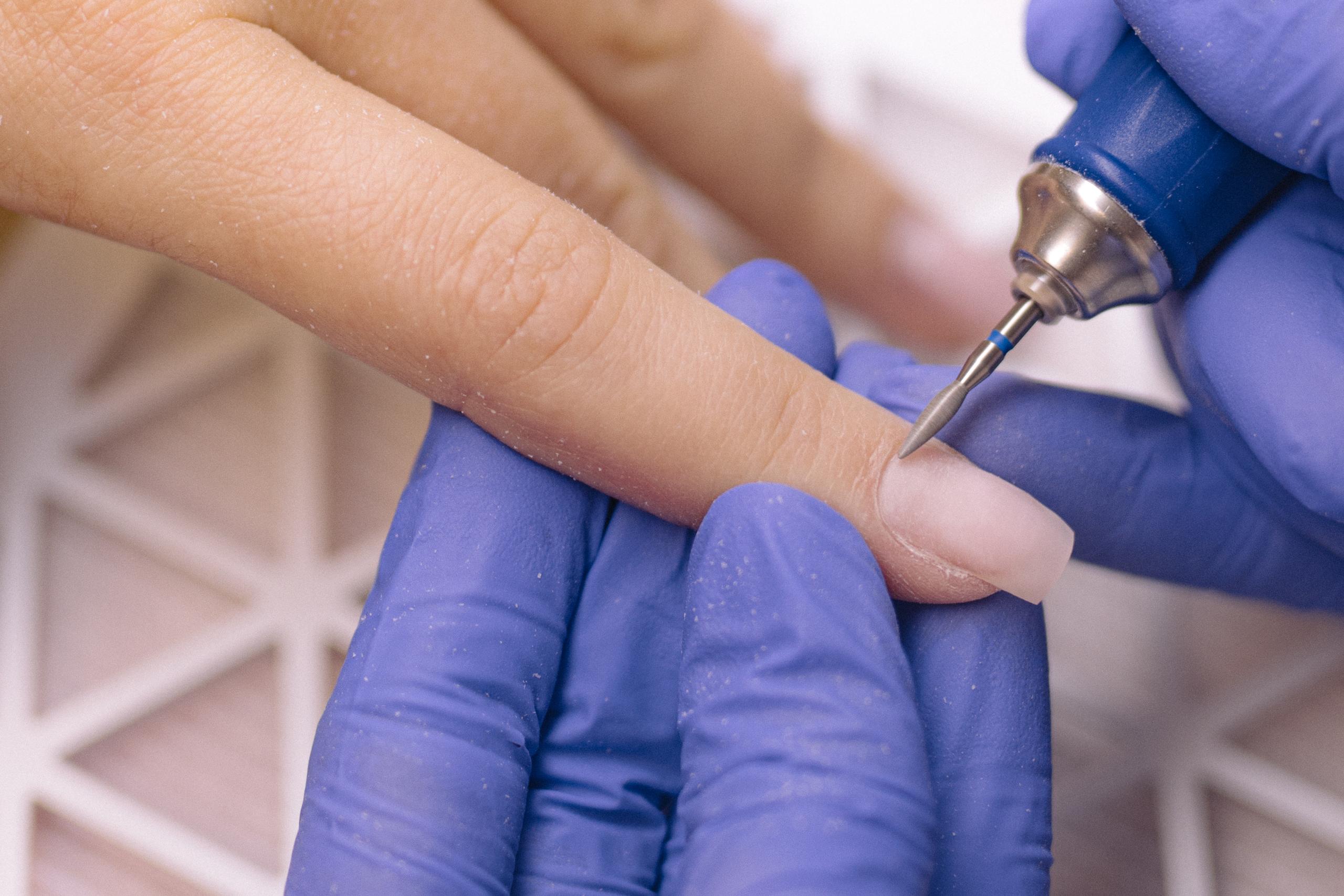 A gloved hand uses an electric nail file to shape an artificial nail.
