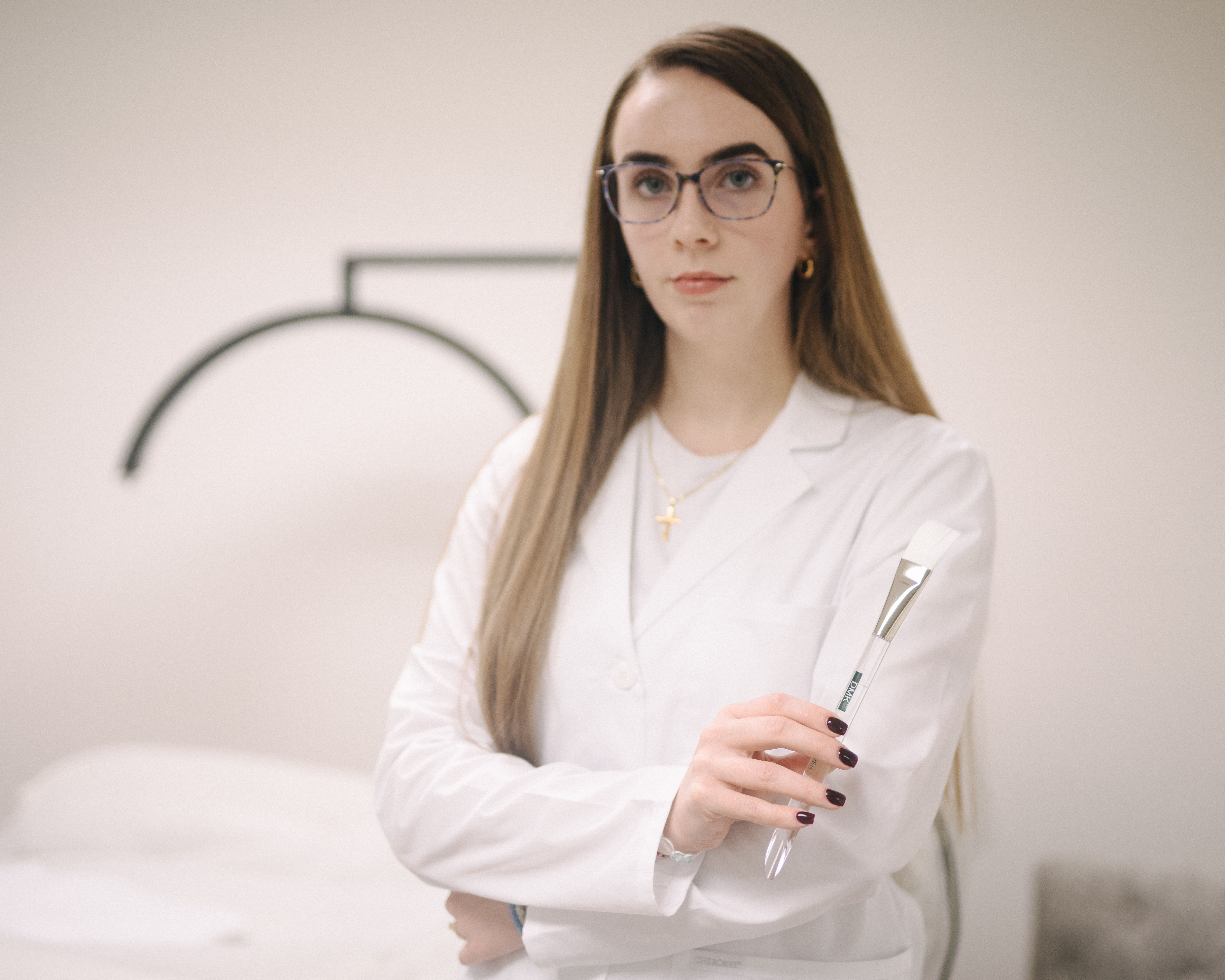 An esthetician in a white lab coat and glasses holds a facial brush.