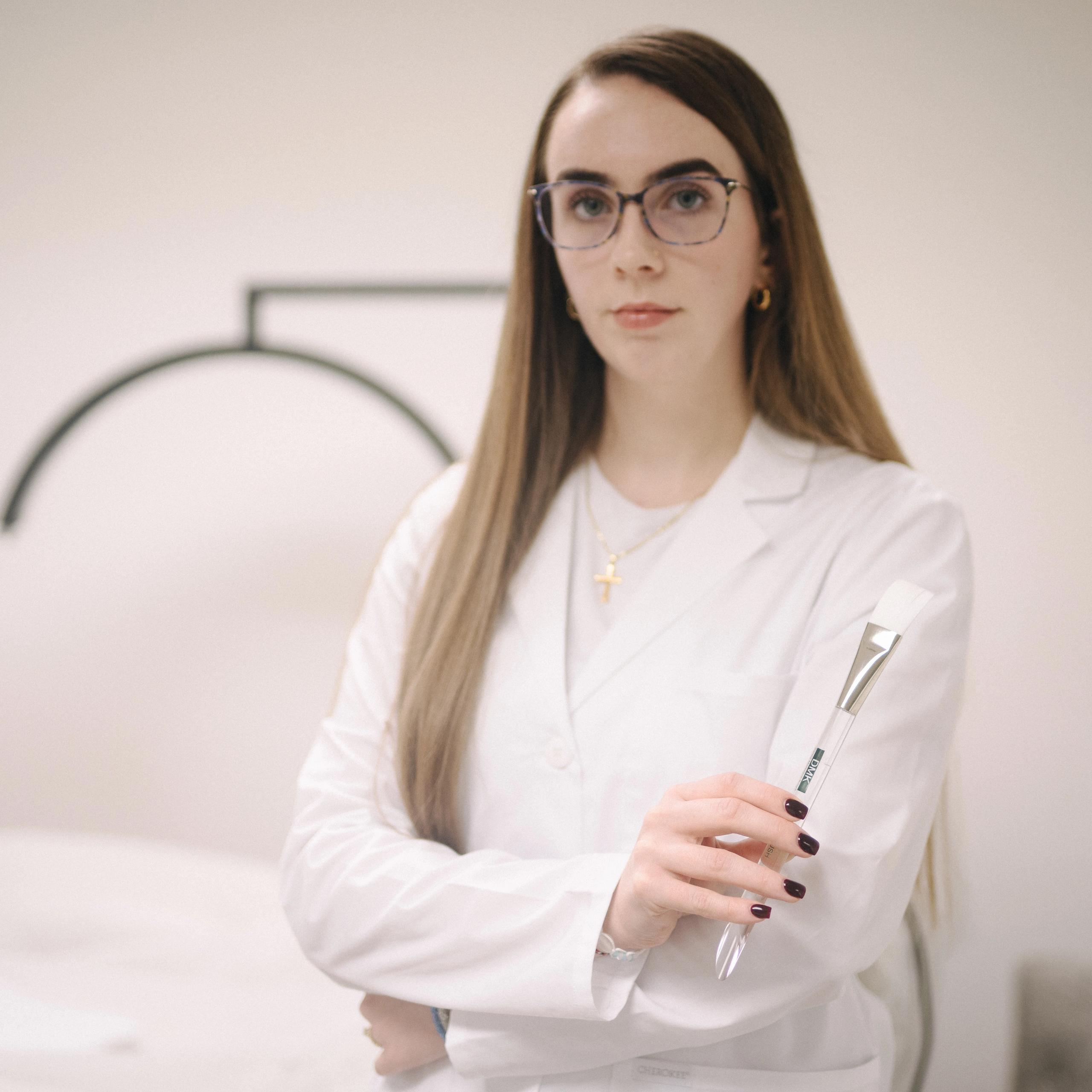 An esthetician in a white lab coat and glasses holds a facial brush.