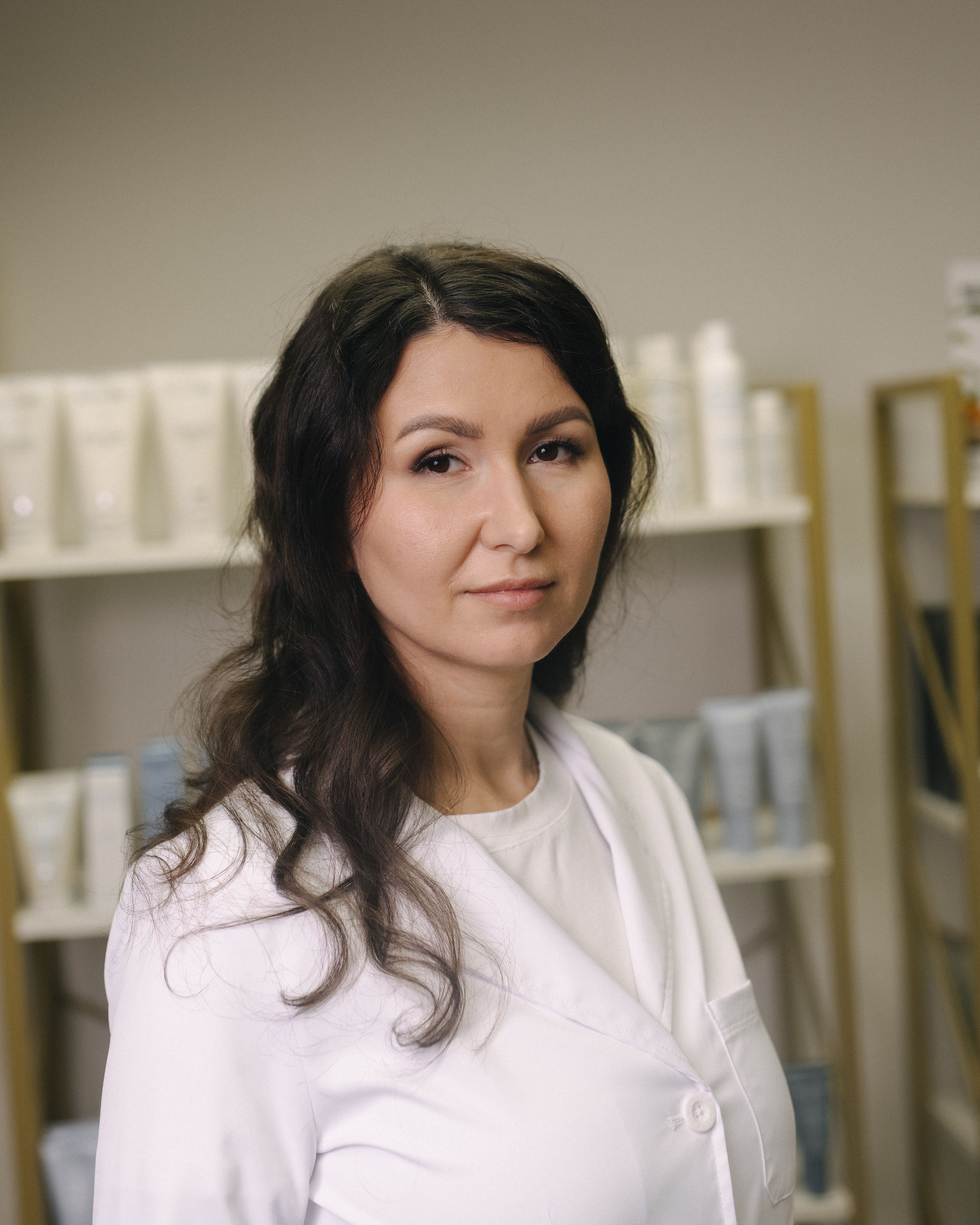 A woman in a white lab coat with dark, wavy hair looks forward in a beauty salon setting.