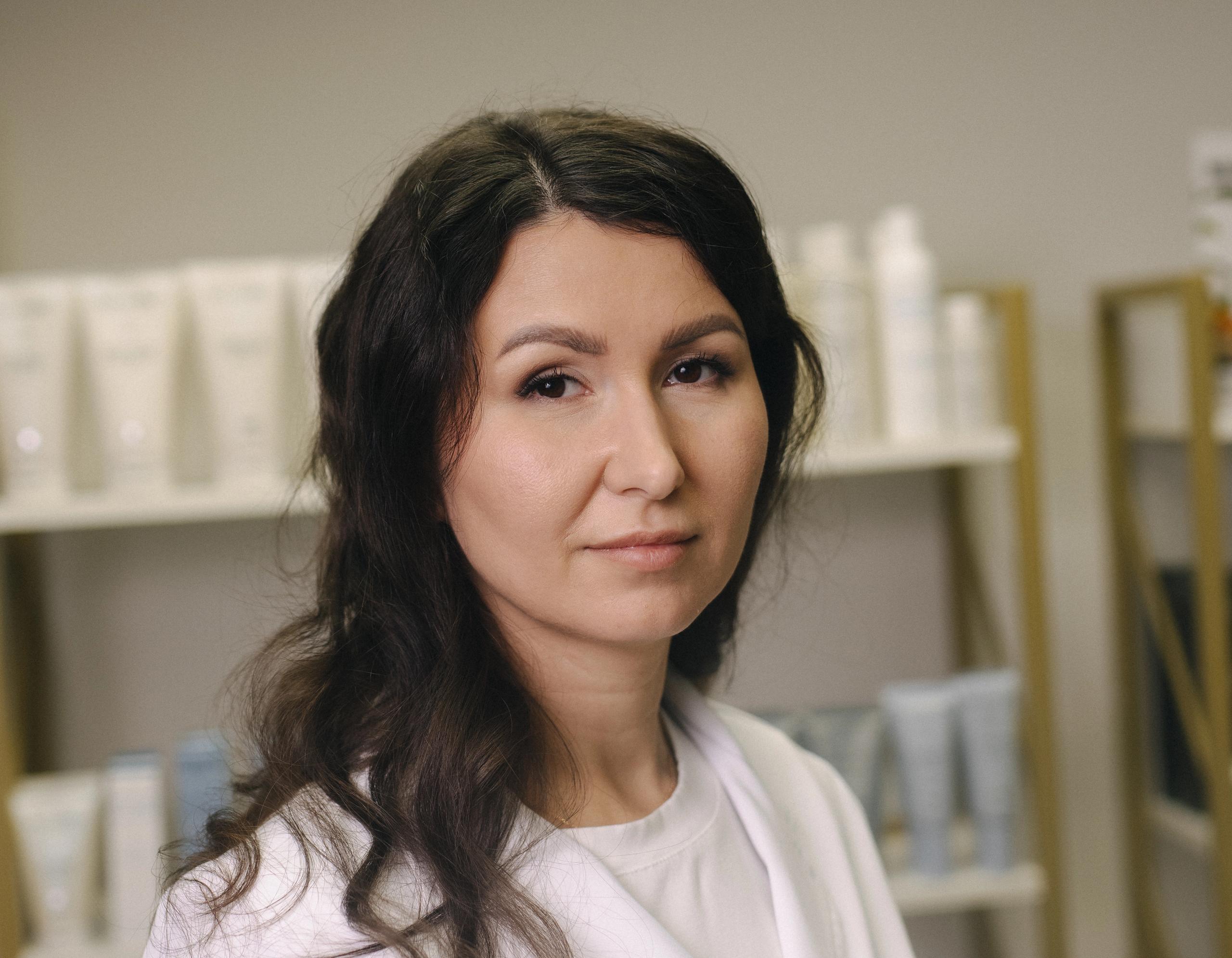 A woman in a white lab coat with dark, wavy hair looks forward in a beauty salon setting.