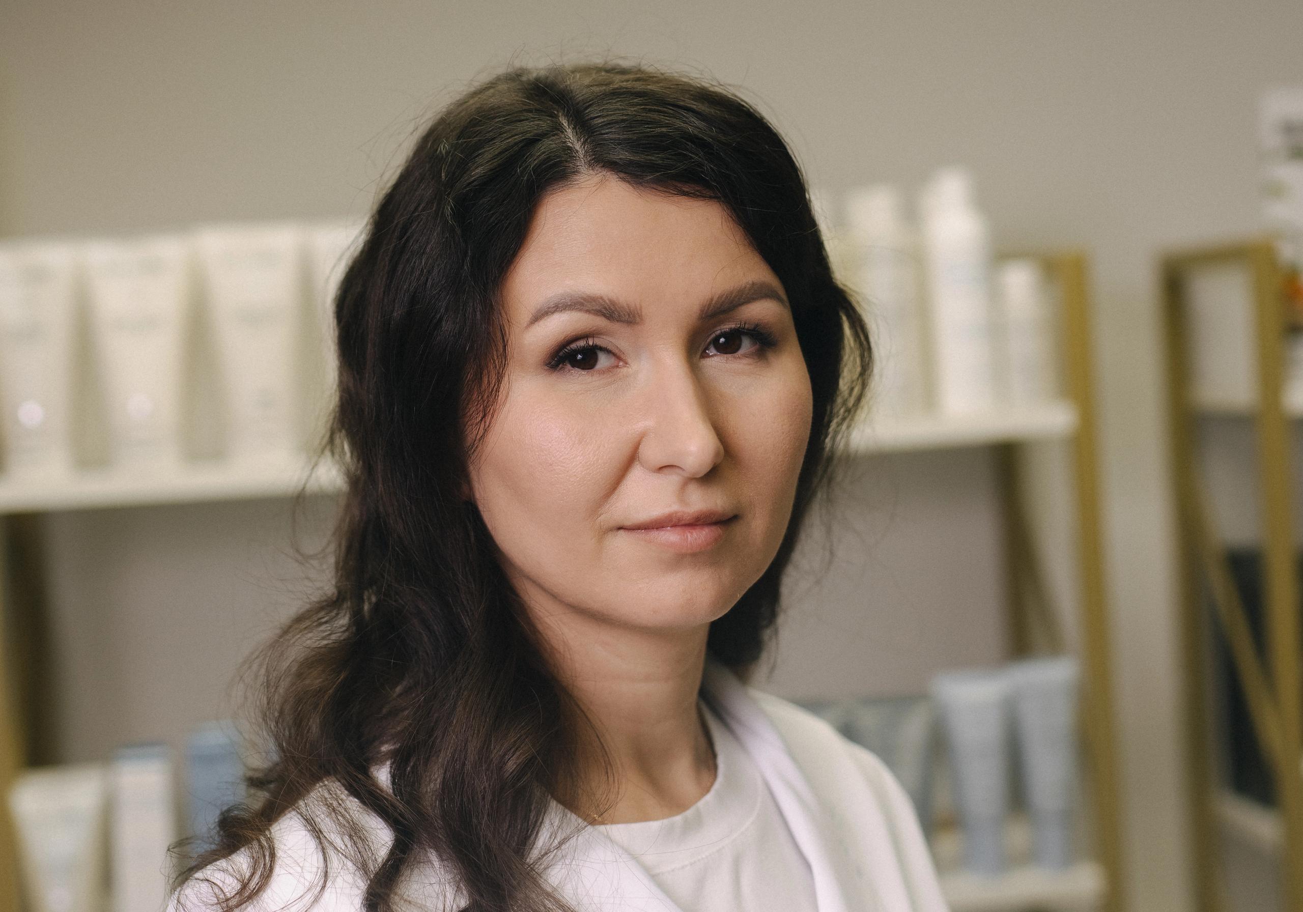A woman in a white lab coat with dark, wavy hair looks forward in a beauty salon setting.