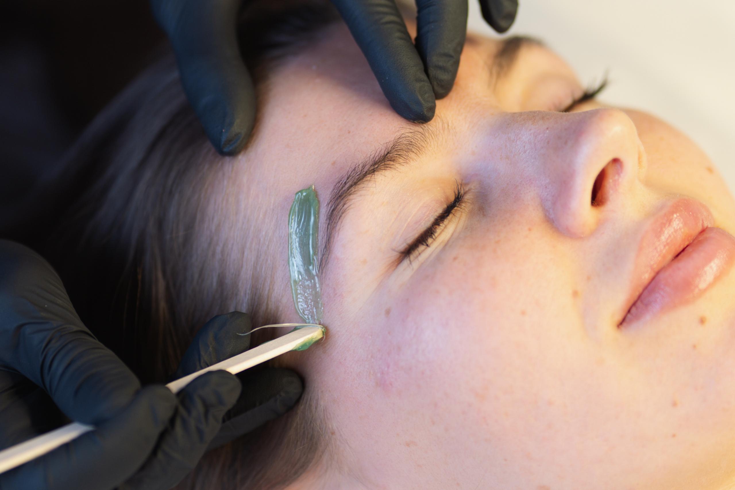 A person applies green wax to a woman's eyebrow for waxing.