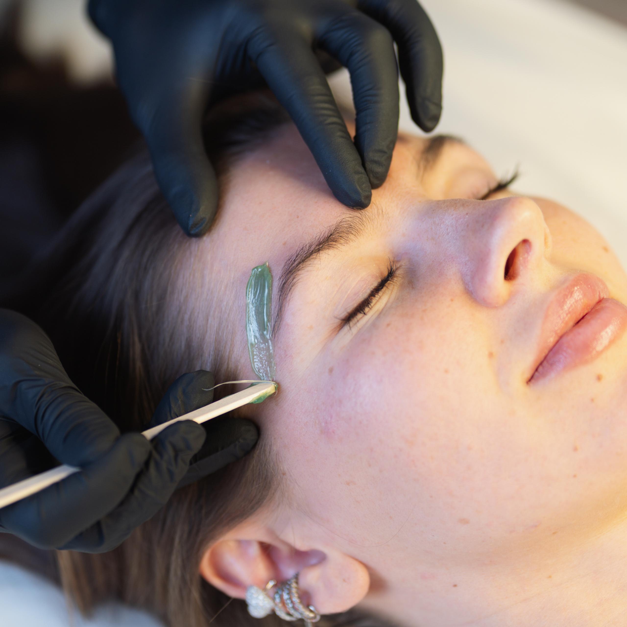 Close-up of a woman getting her eyebrow waxed by gloved hands.