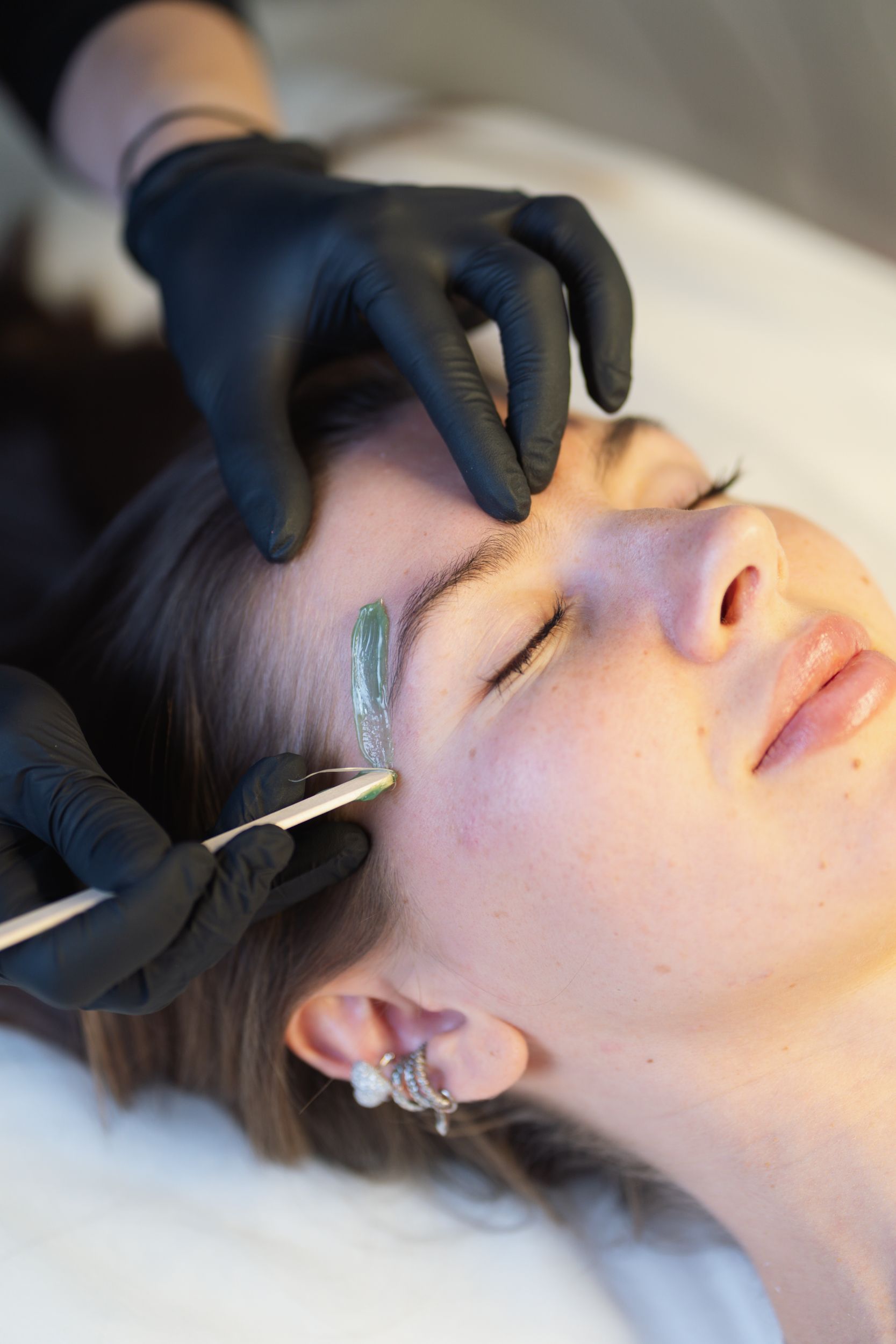Gloved esthetician applying green wax beneath a client's eyebrow with a wooden spatula.