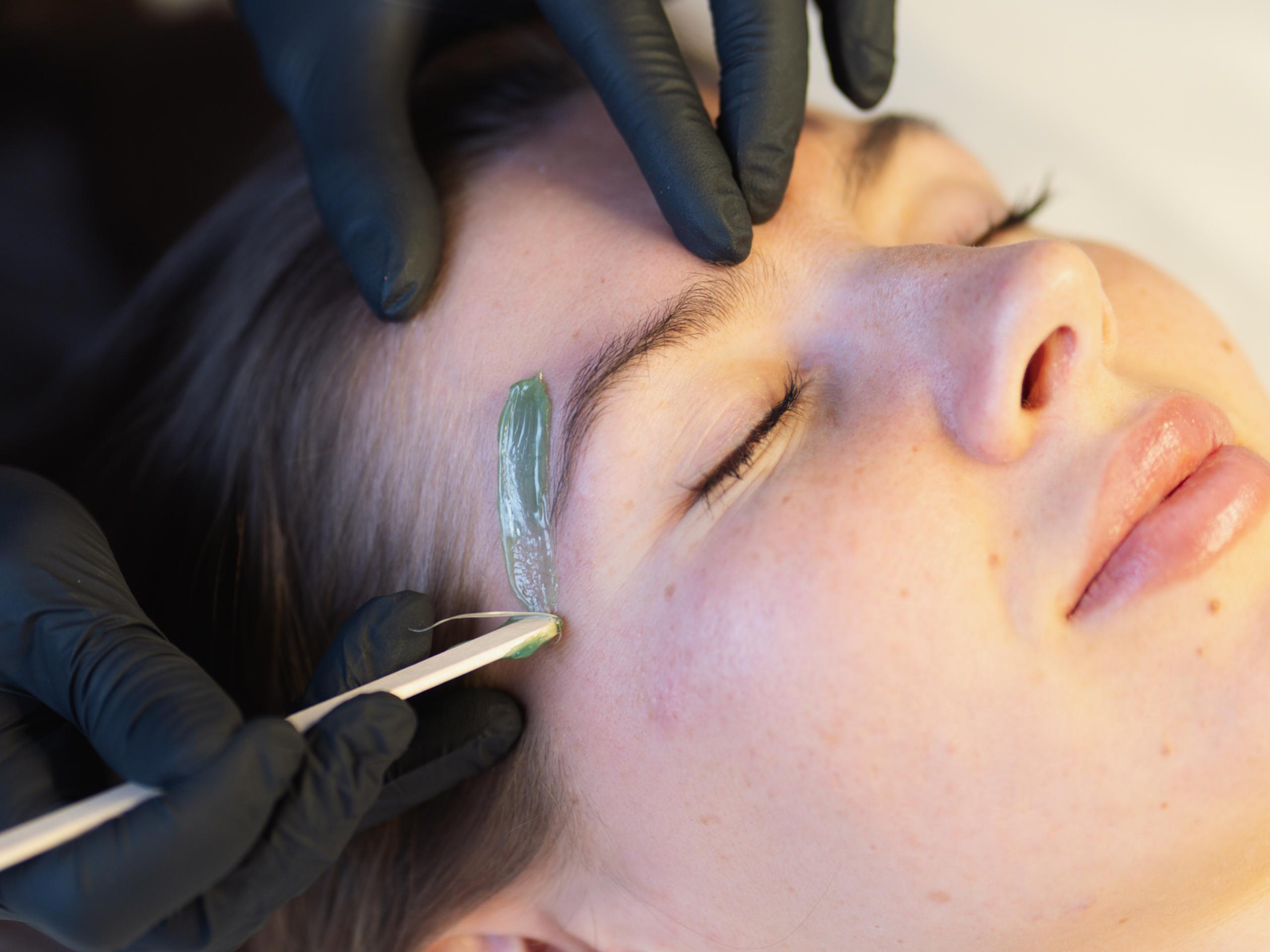 Gloved esthetician applying green wax beneath a client's eyebrow with a wooden spatula.