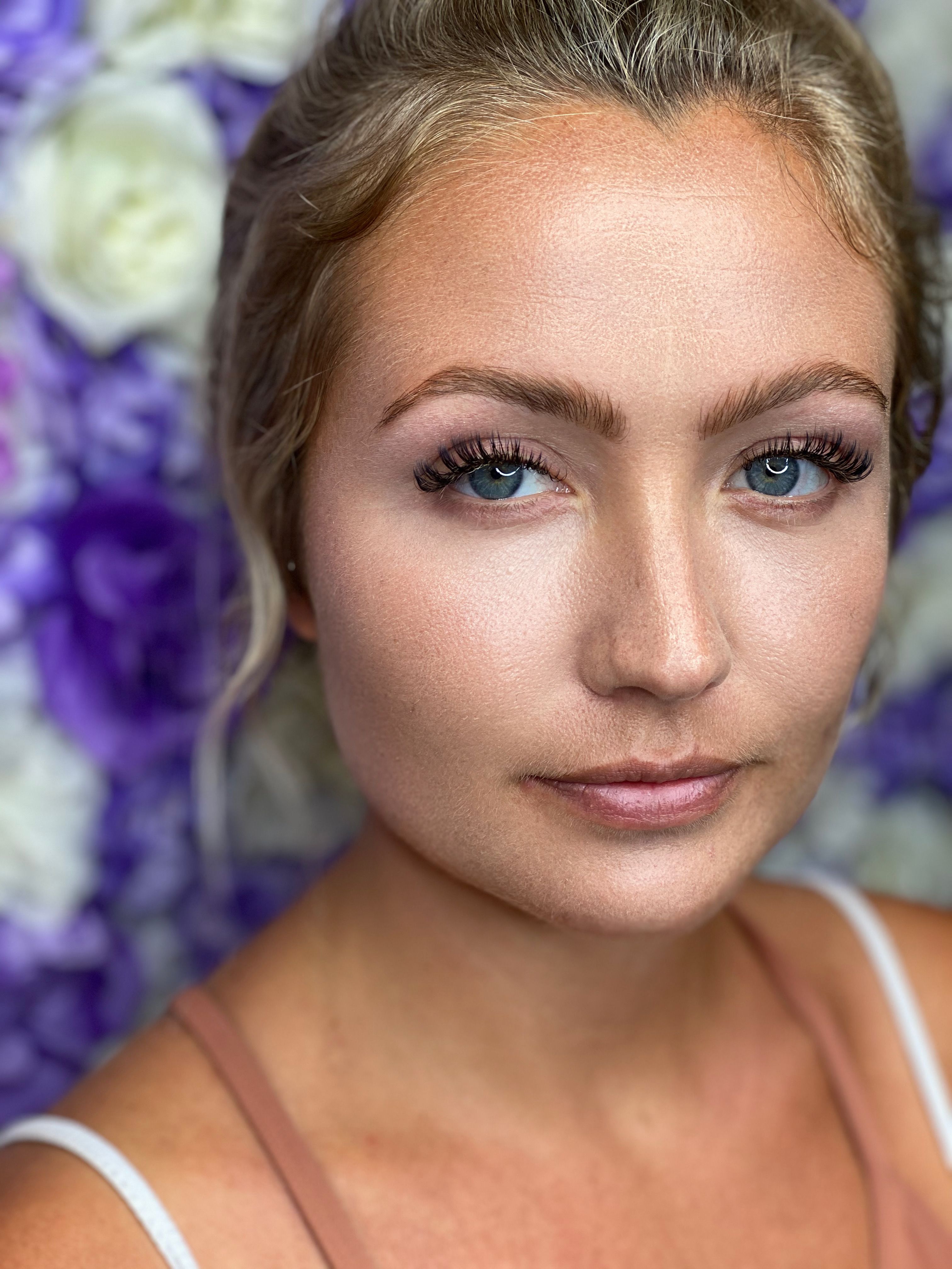 A blonde woman with blue eyes and long eyelashes against a blurred purple and floral background.