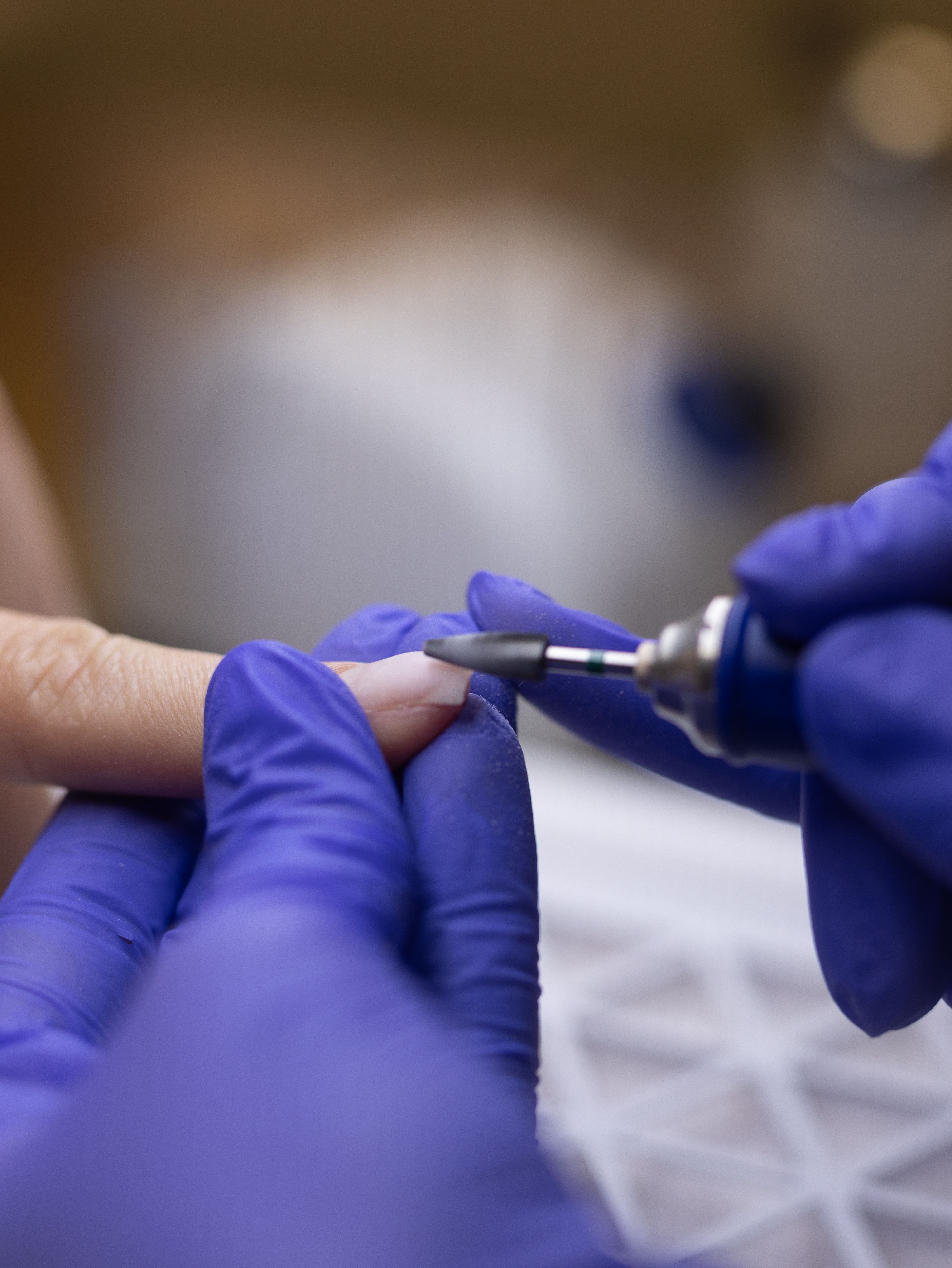 A close-up of a person in purple gloves using a nail drill to shape a fingernail.