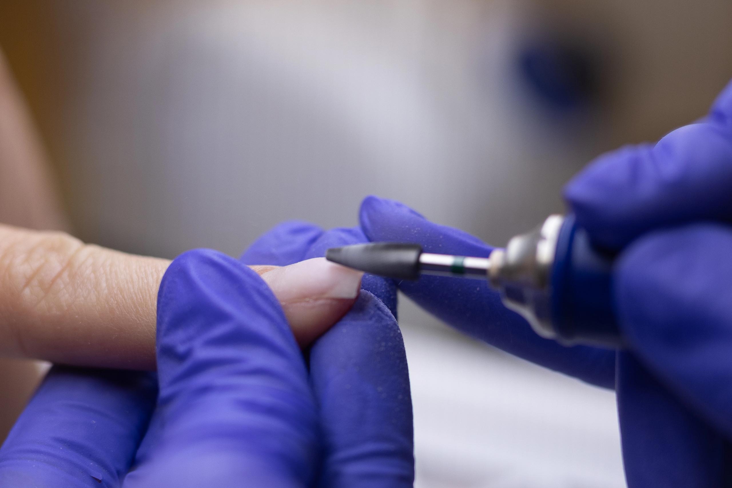 A close-up of a person in purple gloves using a nail drill to shape a fingernail.