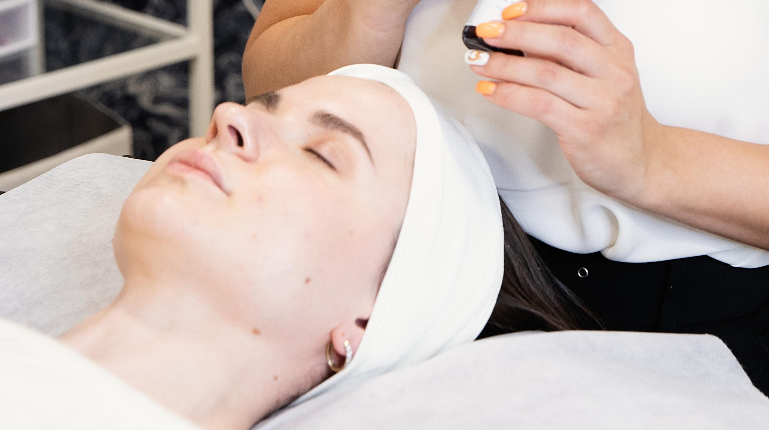 A skincare aesthetician looking down at a client lying on a spa bed.