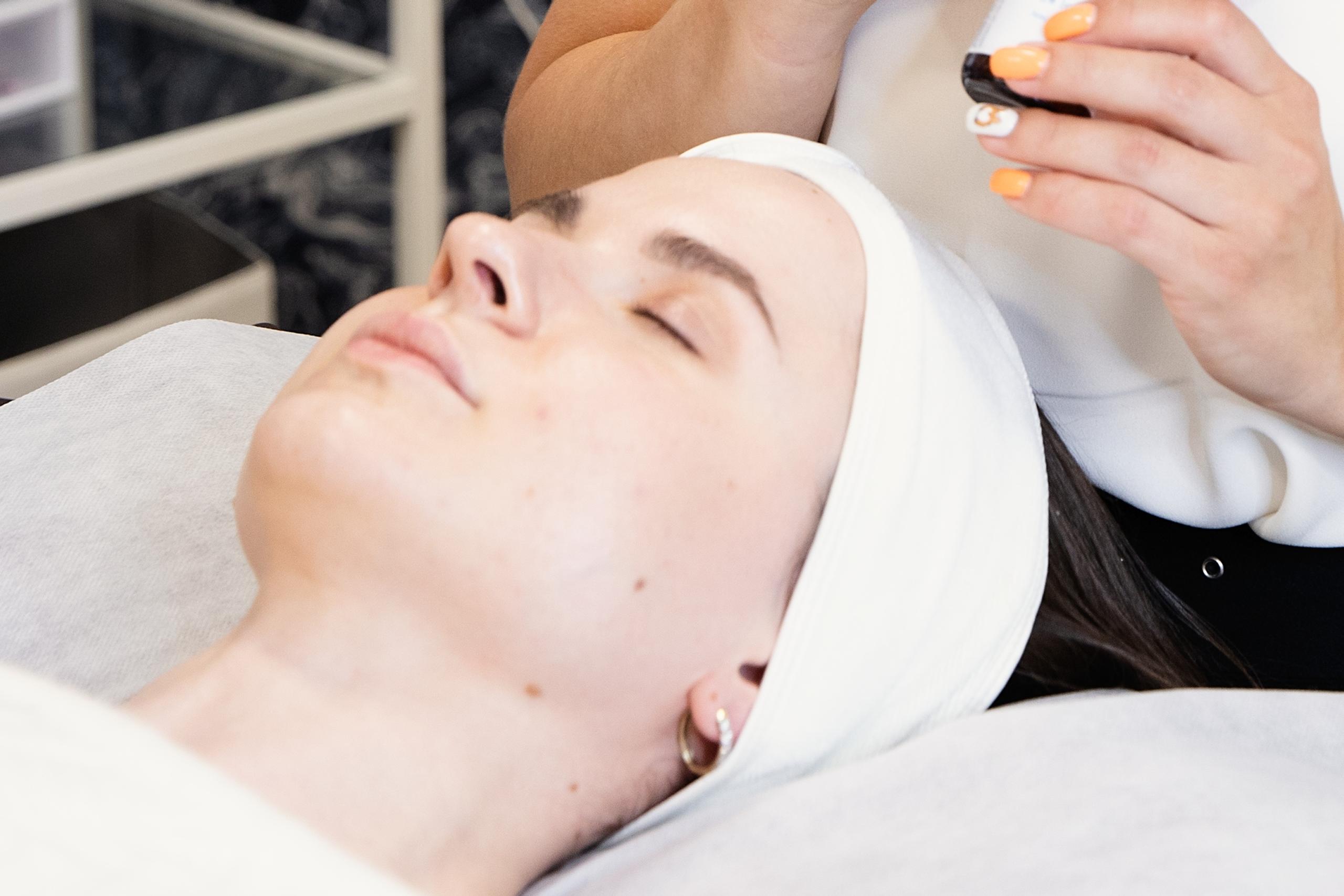 A skincare aesthetician looking down at a client lying on a spa bed.