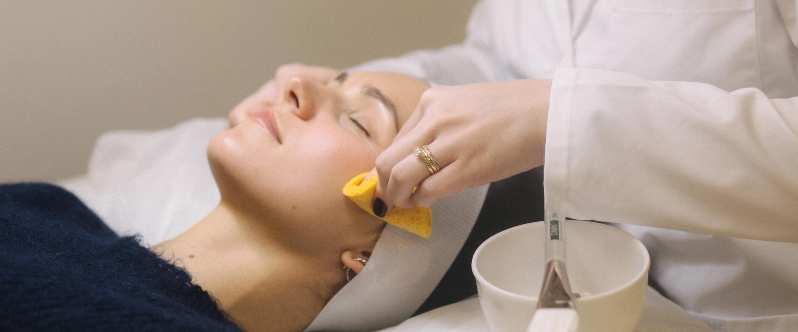 A woman receives a facial treatment, an aesthetician cleansing her cheek with a sponge.