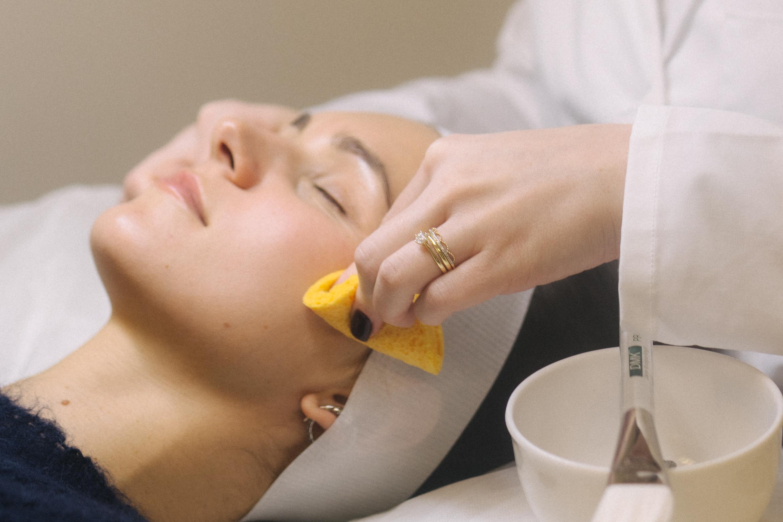 A woman receives a facial treatment, an aesthetician cleansing her cheek with a sponge.