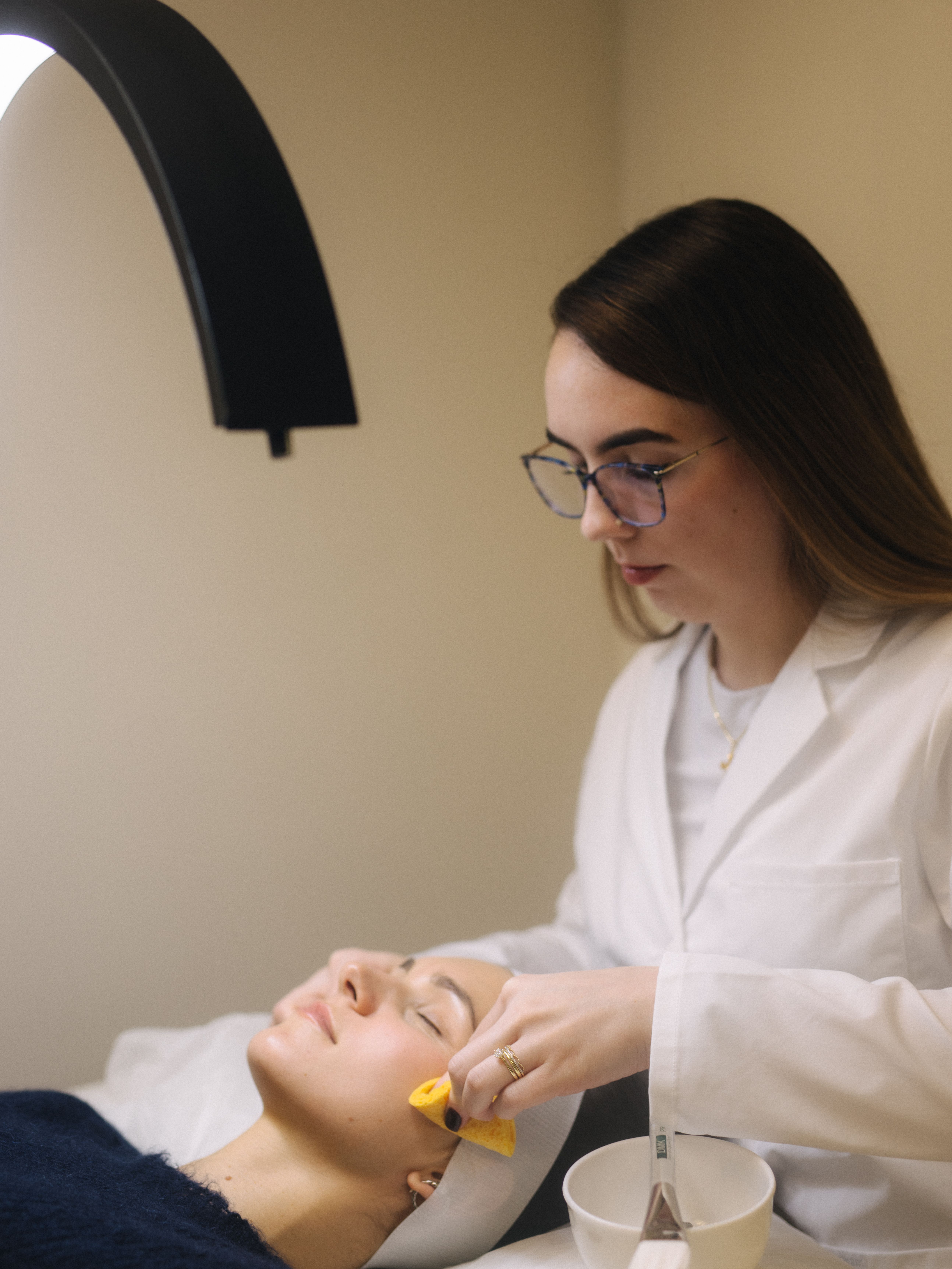 An aesthetician in a white lab coat wipes a client's cheek with a yellow sponge during a facial.