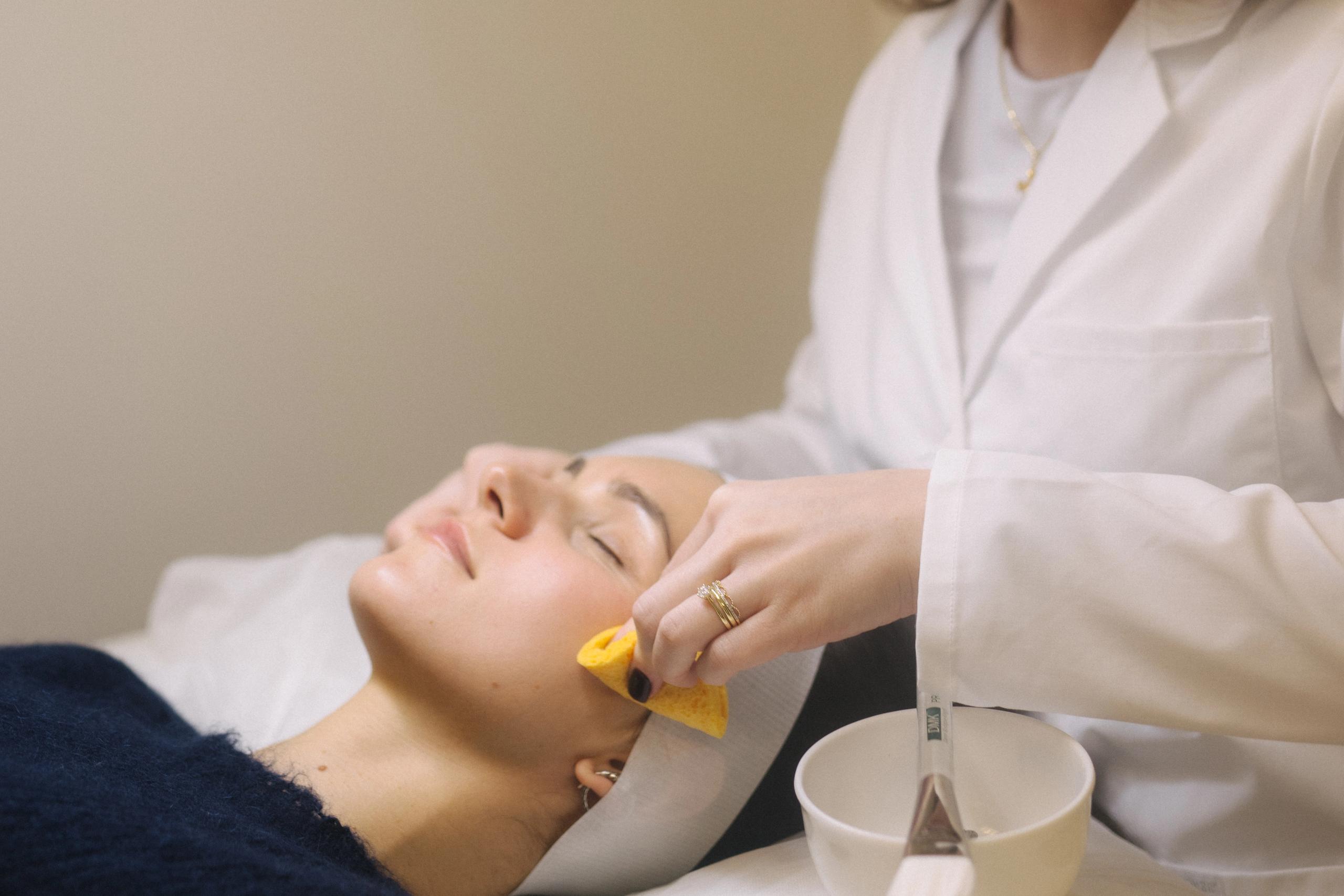 An aesthetician in a white lab coat wipes a client's cheek with a yellow sponge during a facial.