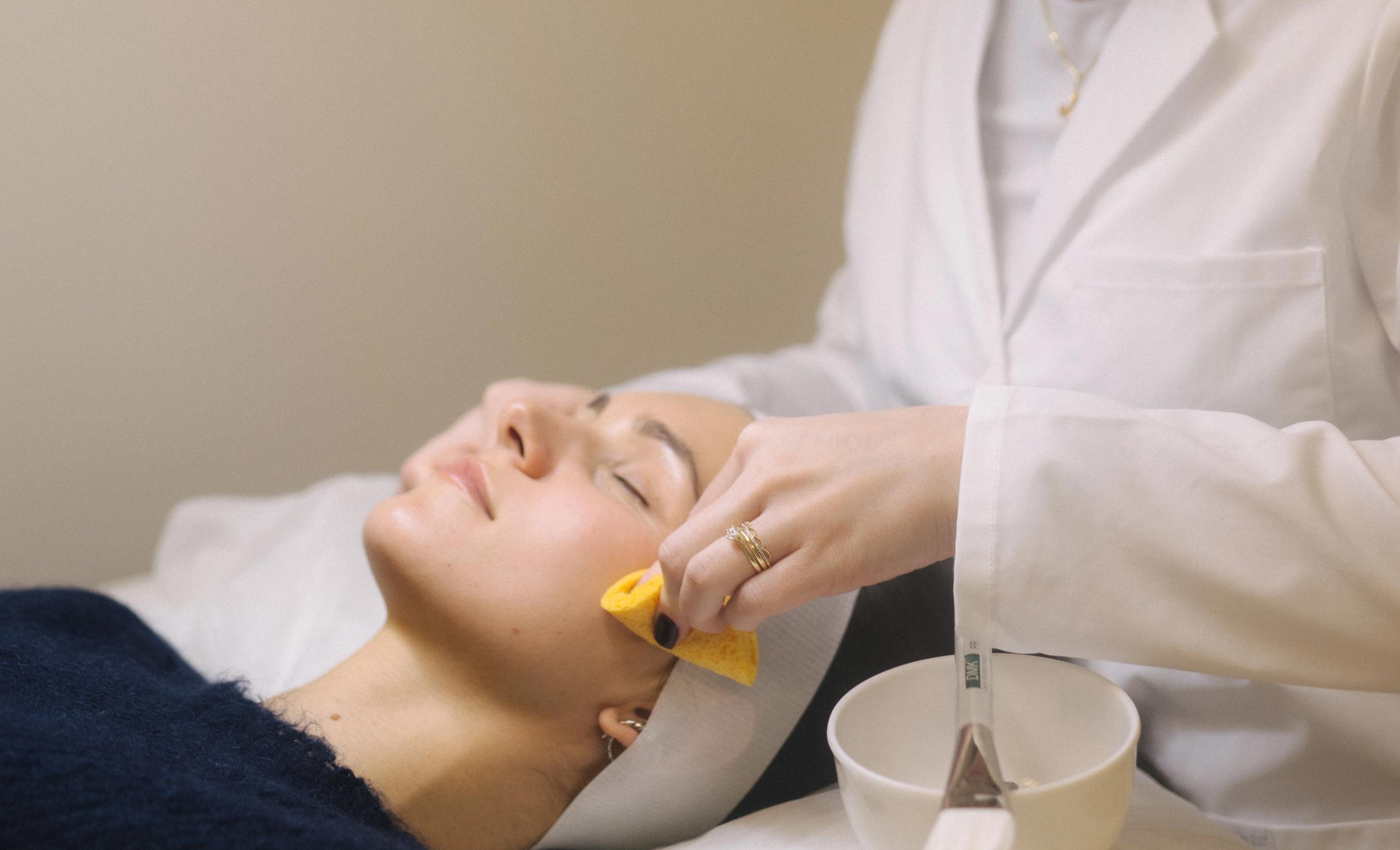 An aesthetician in a white lab coat wipes a client's cheek with a yellow sponge during a facial.