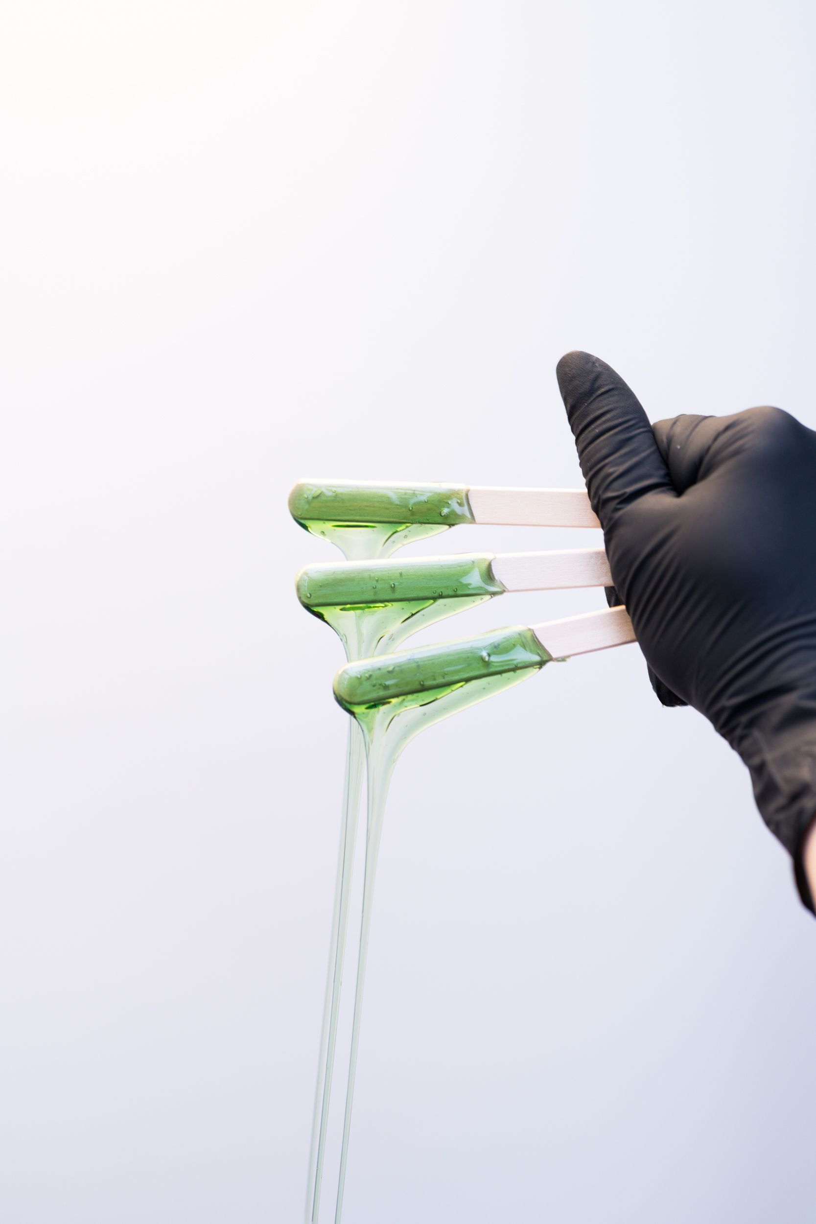 Close-up of a hand in black gloves holding wooden spatulas coated with green wax dripping.