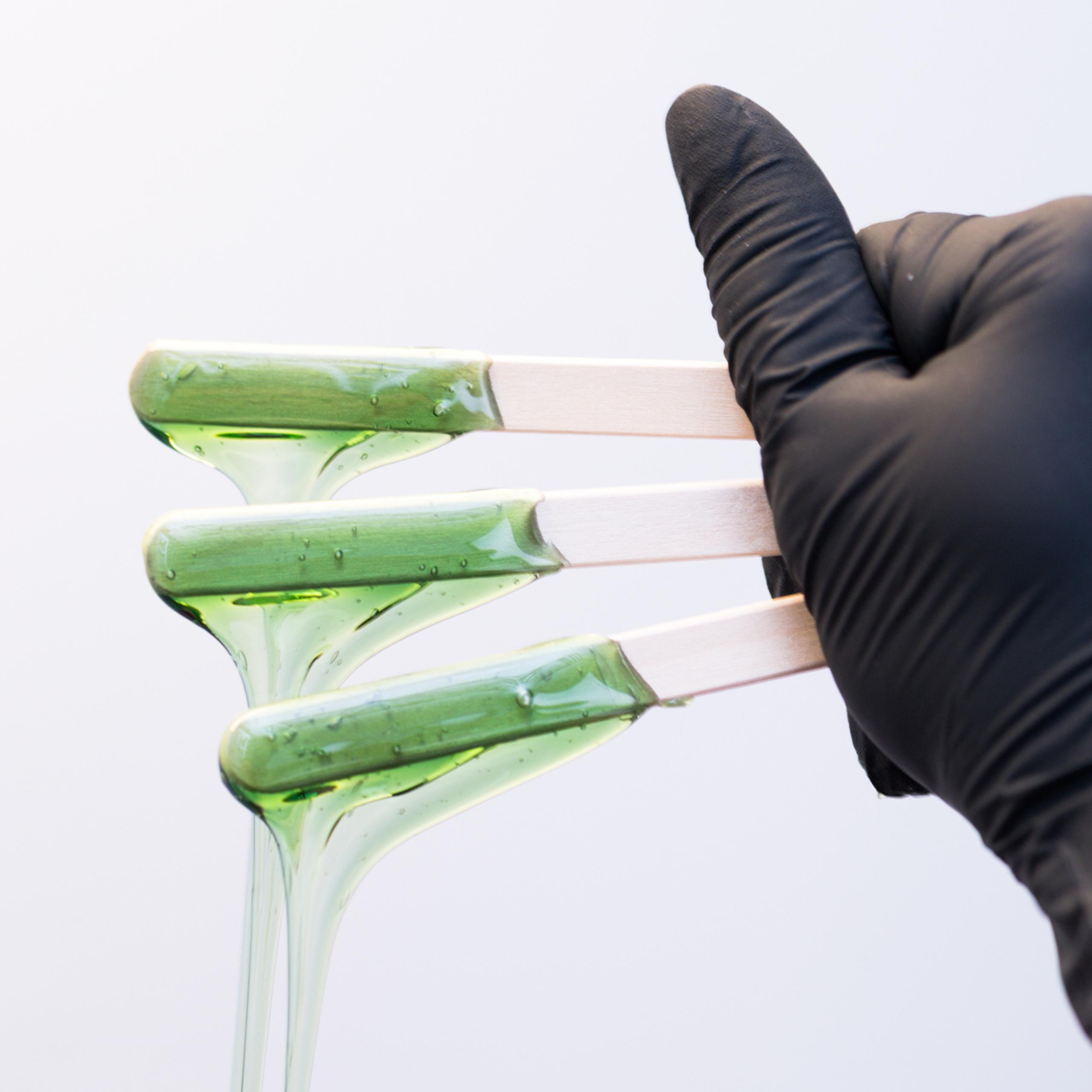 Close-up of a hand in black gloves holding wooden spatulas coated with green wax dripping.