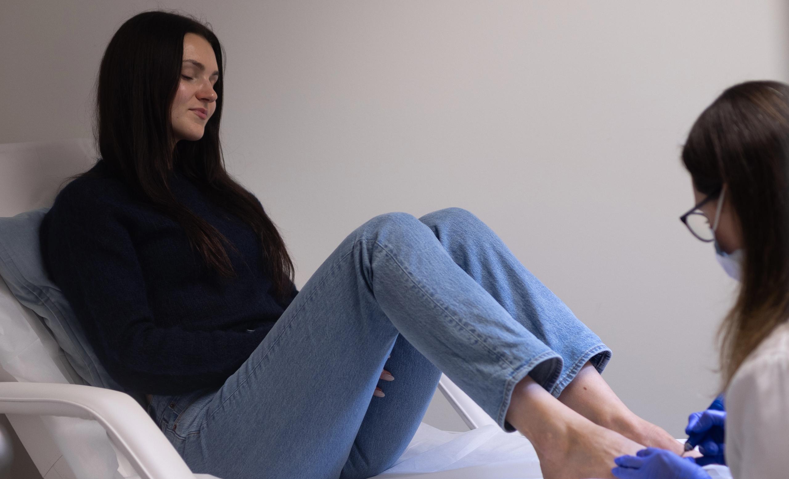 A woman on a white treatment bed receives a pedicure from a gloved technician.