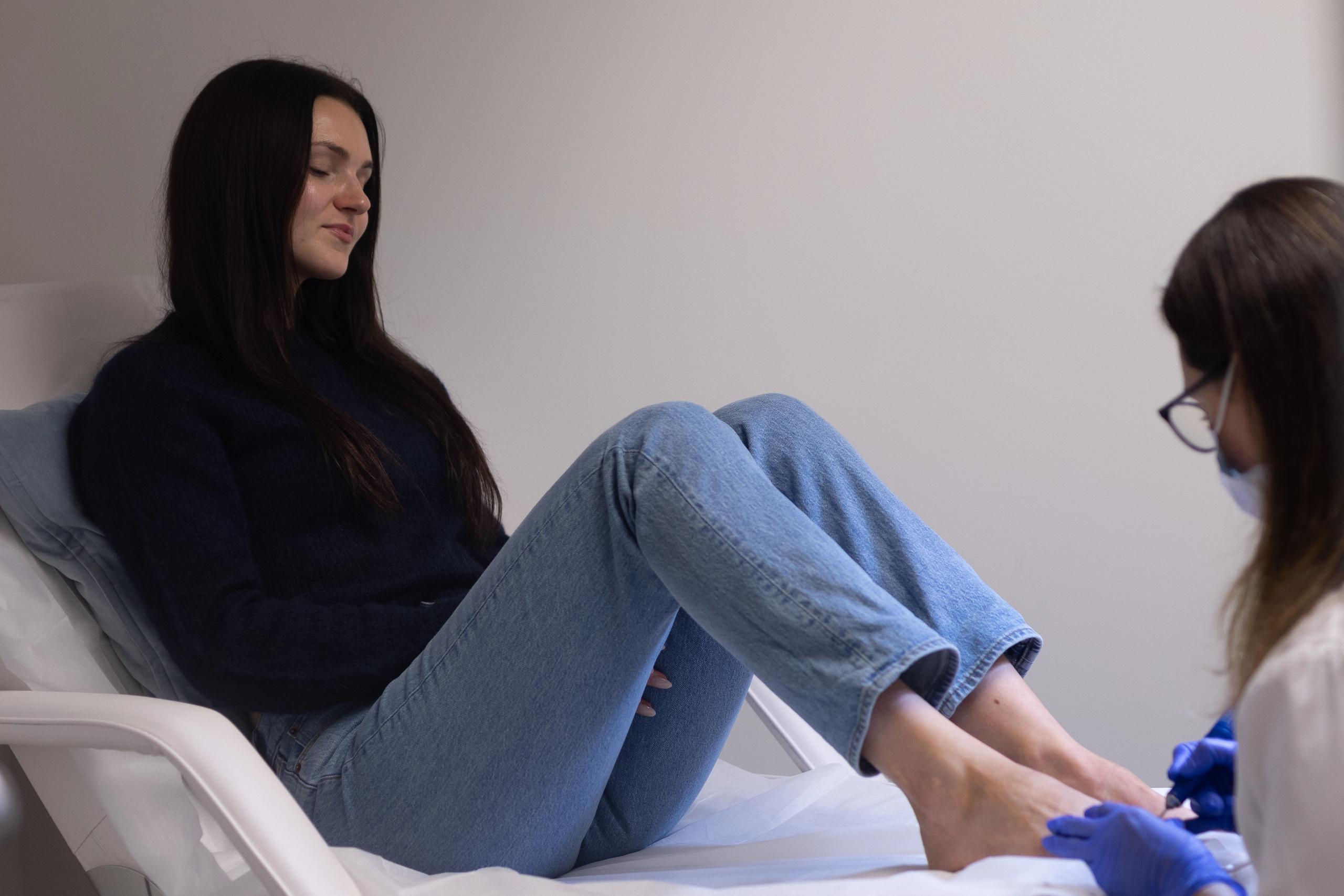 A woman on a white treatment bed receives a pedicure from a gloved technician.