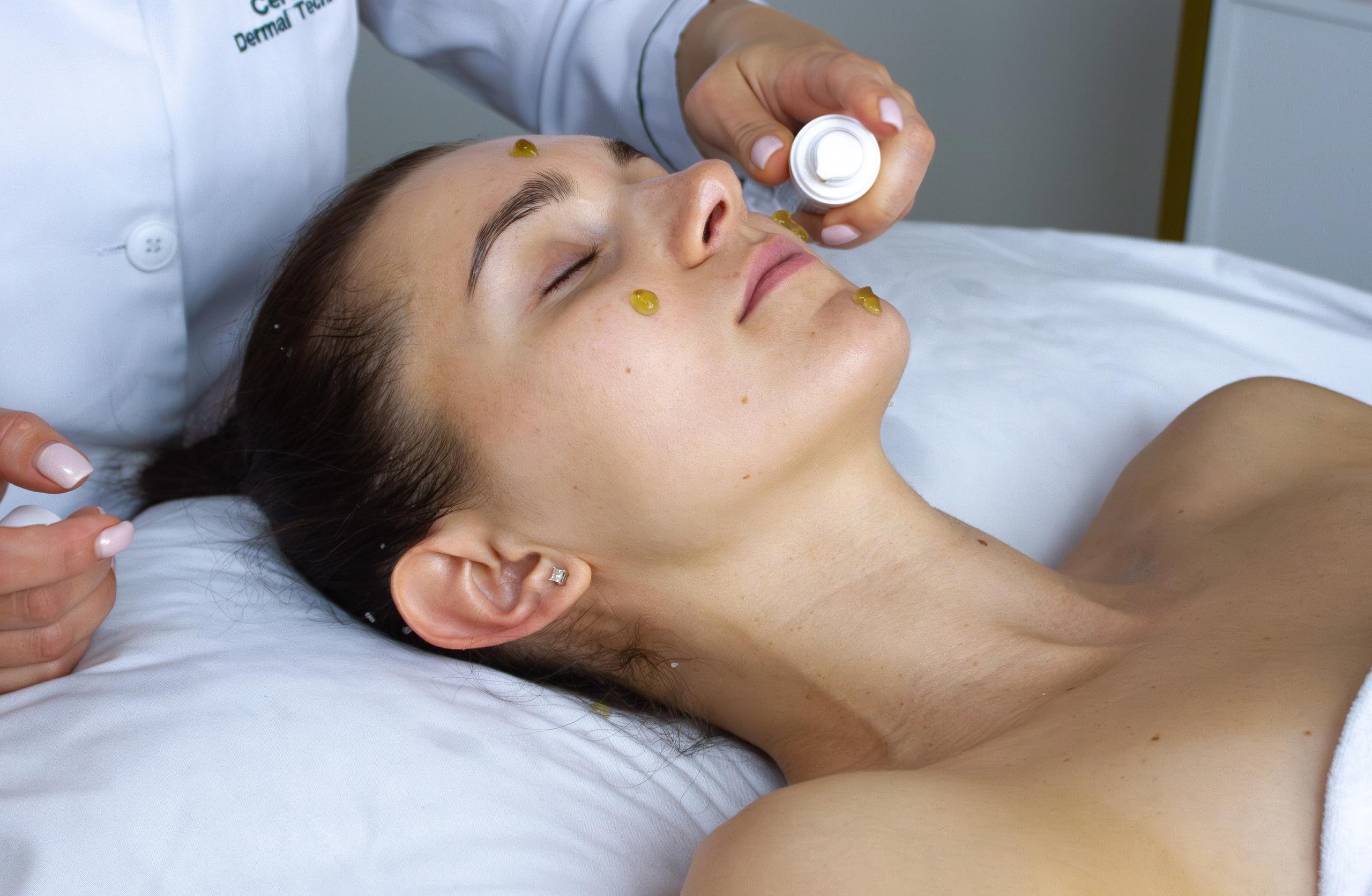A woman lies with her eyes closed as a technician applies product dots to her face.