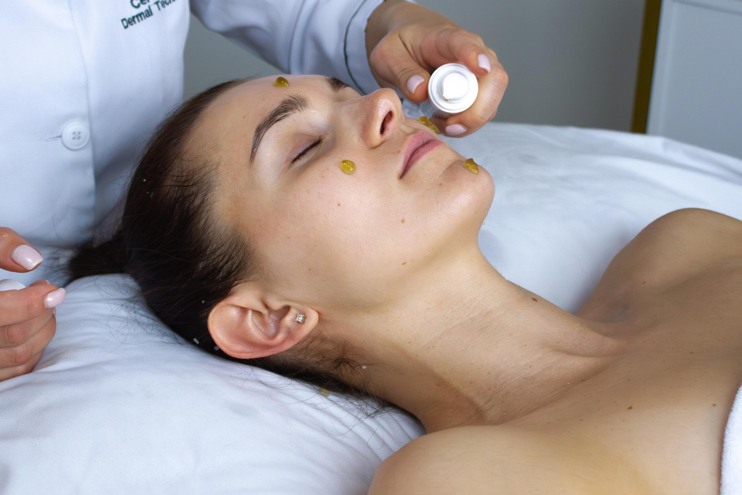 A woman lies with her eyes closed as a technician applies product dots to her face.