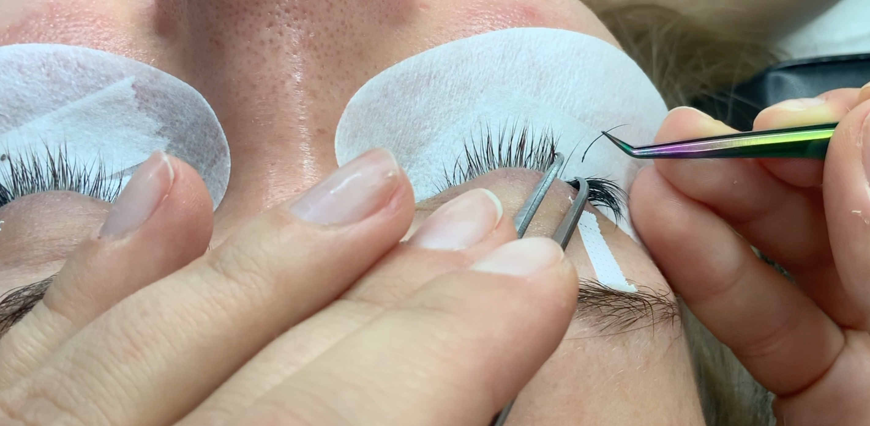 Lash artist applying eyelash extensions with tweezers while protective pads cover the client's under-eye area