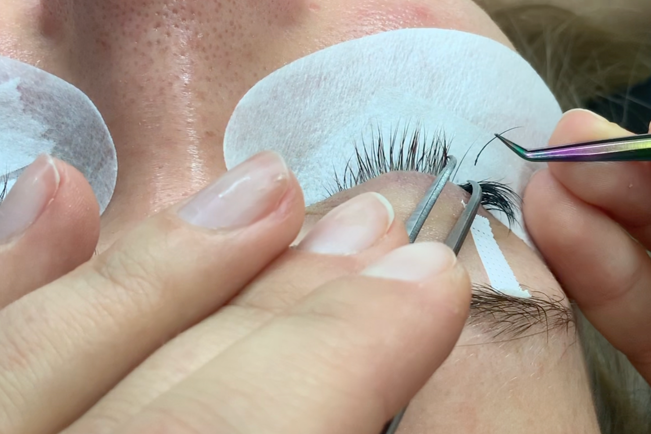 Lash artist applying eyelash extensions with tweezers while protective pads cover the client's under-eye area