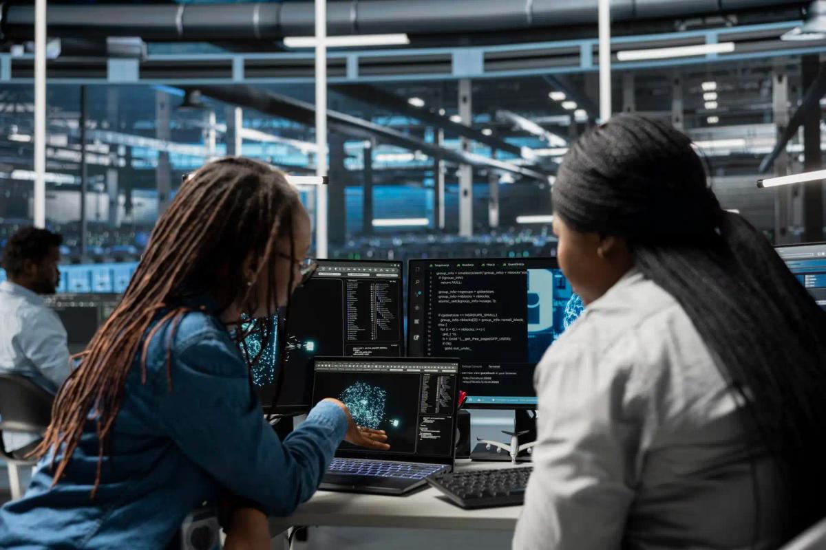 Foto de duas profissionais de tecnologia colaborando em testes de automação, analisando códigos e gráficos em múltiplos monitores dentro de um ambiente moderno de QA.