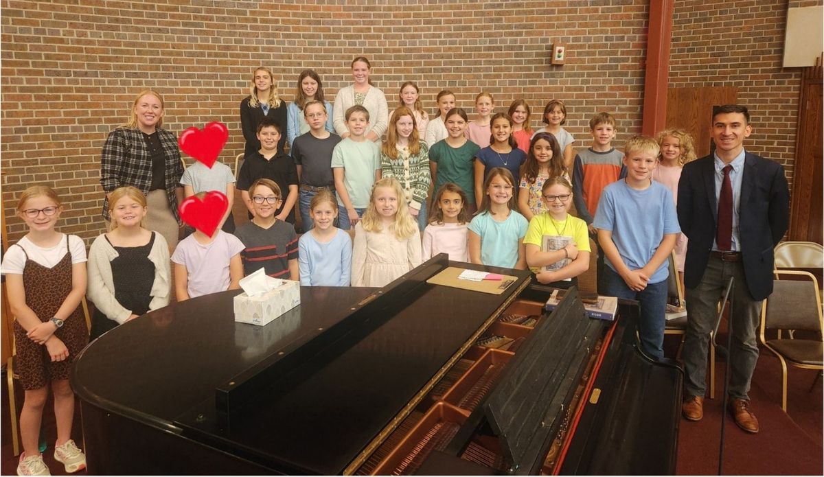 St. Aloysius School choir and teachers gathered around a piano; two faces retain the newsletter’s red-heart coverings